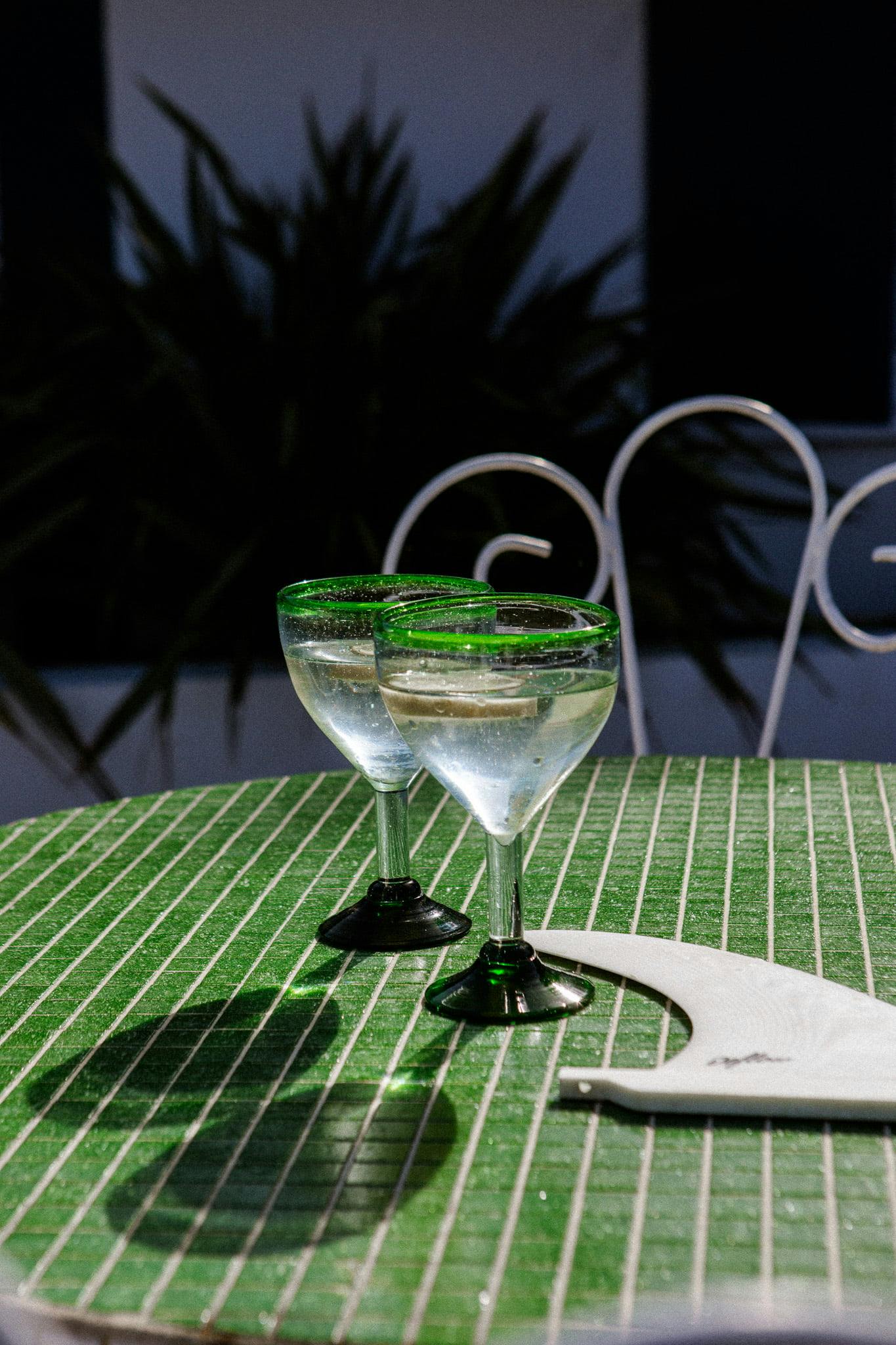 Deux verres à margarita aux bords verts sont posés sur une table verte à rayures. Les verres semblent remplis d’un liquide clair. Une serviette blanche est placée à côté des verres. L'arrière-plan présente une verdure floue et une partie d'une chaise en métal blanc, créant une scène invitante pour un jeu Domino Un.