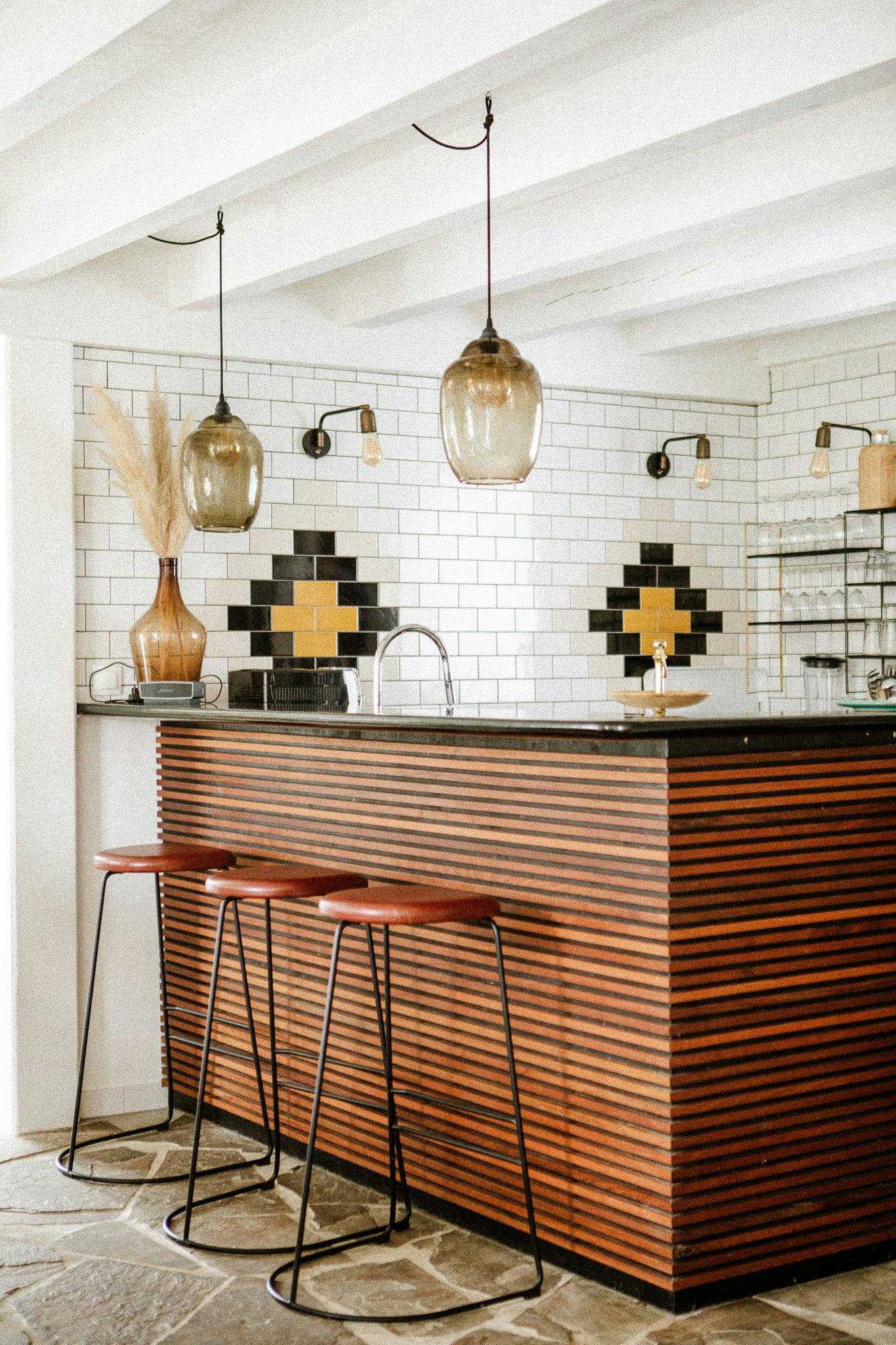 A modern kitchen features a wooden island with two bar stools, pendant lights, and a minimalist backsplash of white tiles accented with black and yellow diamond patterns inspired by Domino Uno. The open and airy space showcases exposed beams, a stone floor, and decorative items like a vase filled with dried grass.