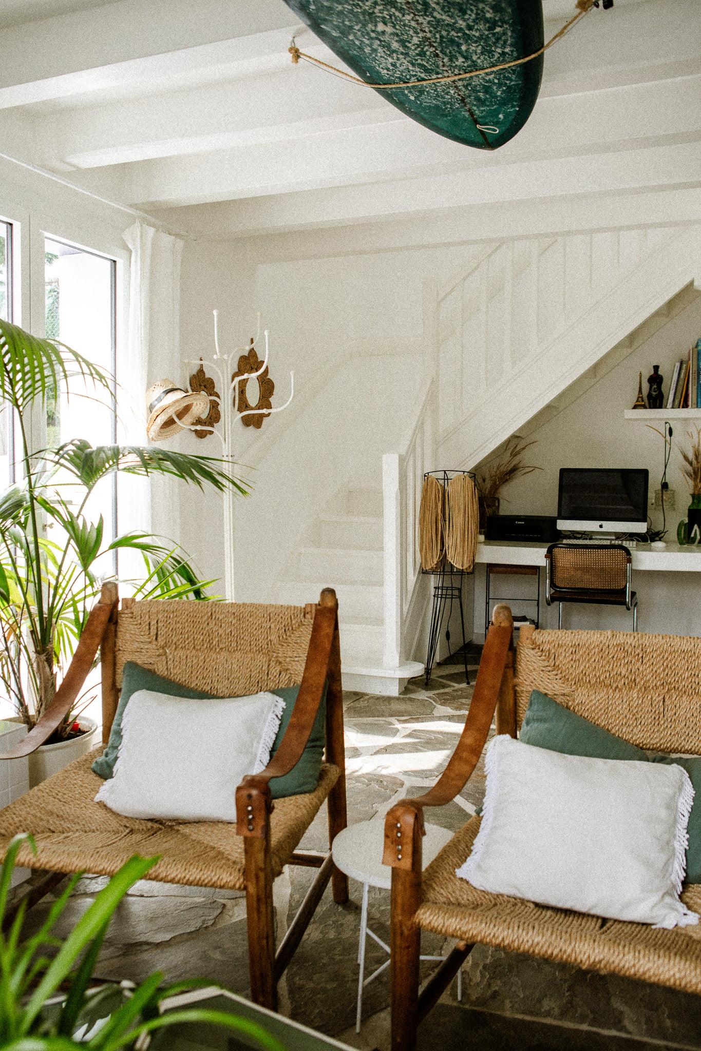 A bright living room features two Domino Uno wicker chairs with white cushions, a small round side table, and an angled desk with a computer. The room is decorated with plants and a surfboard hanging from the ceiling. A staircase leads upstairs, blending seamlessly with the white interior.