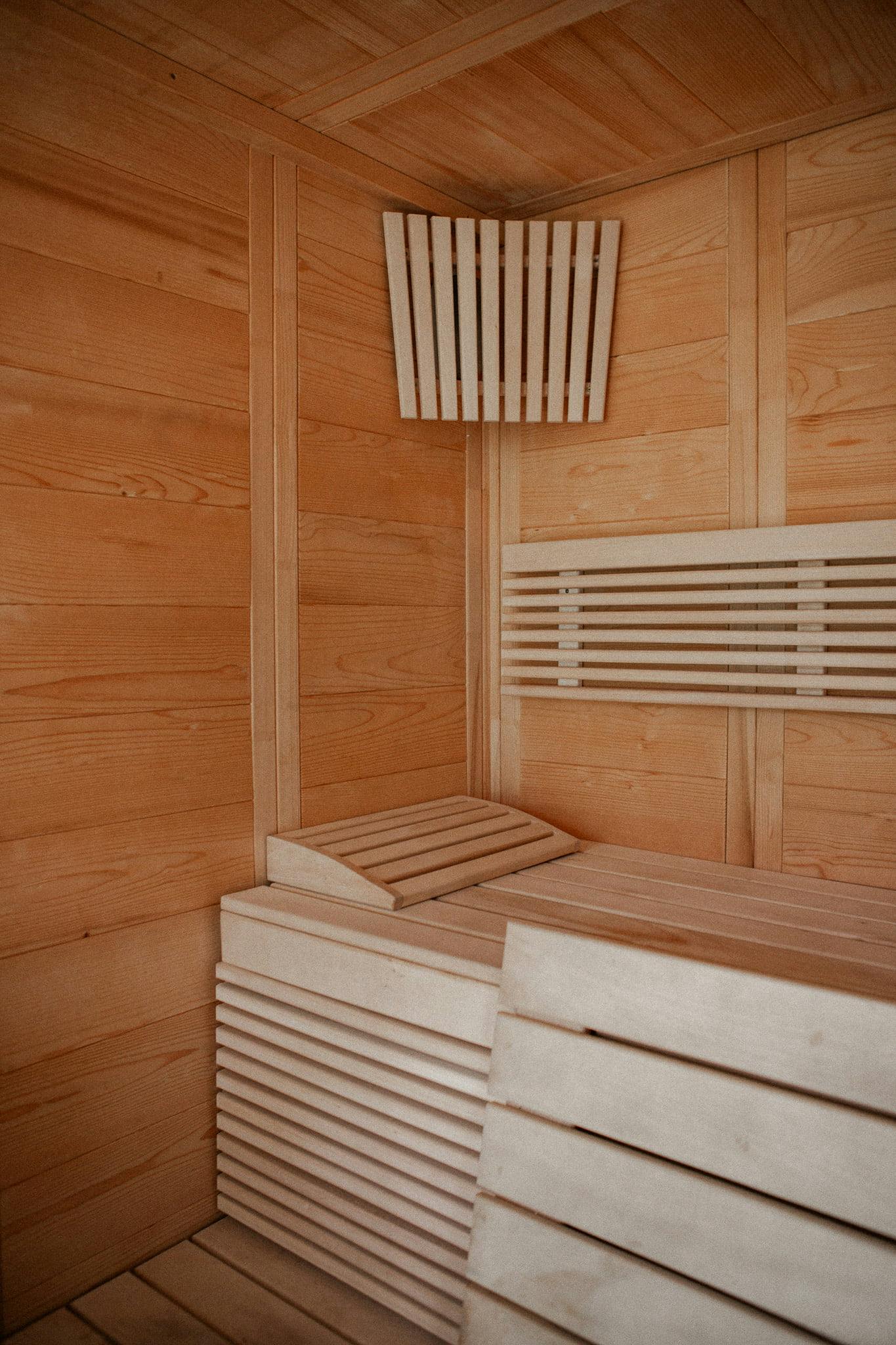 A cozy wooden sauna features light-colored wood paneling covering the walls and ceiling. Wooden benches arranged in tiers provide seating options. This Domino Uno sauna offers a warm and inviting atmosphere, perfect for relaxation.