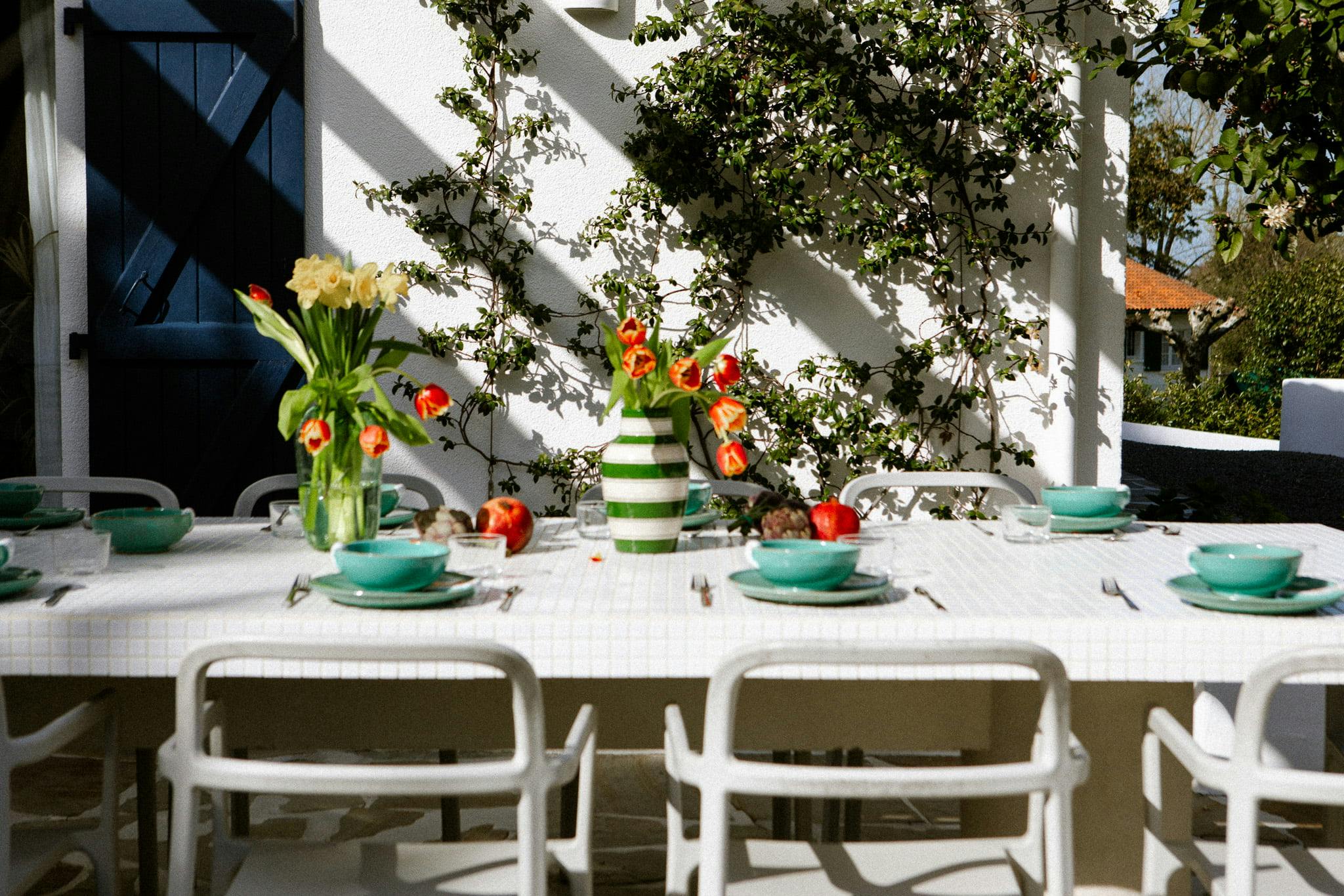 Une table à manger rectangulaire blanche Domino Un est installée à l'extérieur avec des bols verts, des couverts et des pommes rouges. Deux vases à fleurs rouges et jaunes servent de centres de table. L’arrière-plan présente un mur blanc avec une plante grimpante et des ombres d’arbres ou de structures.