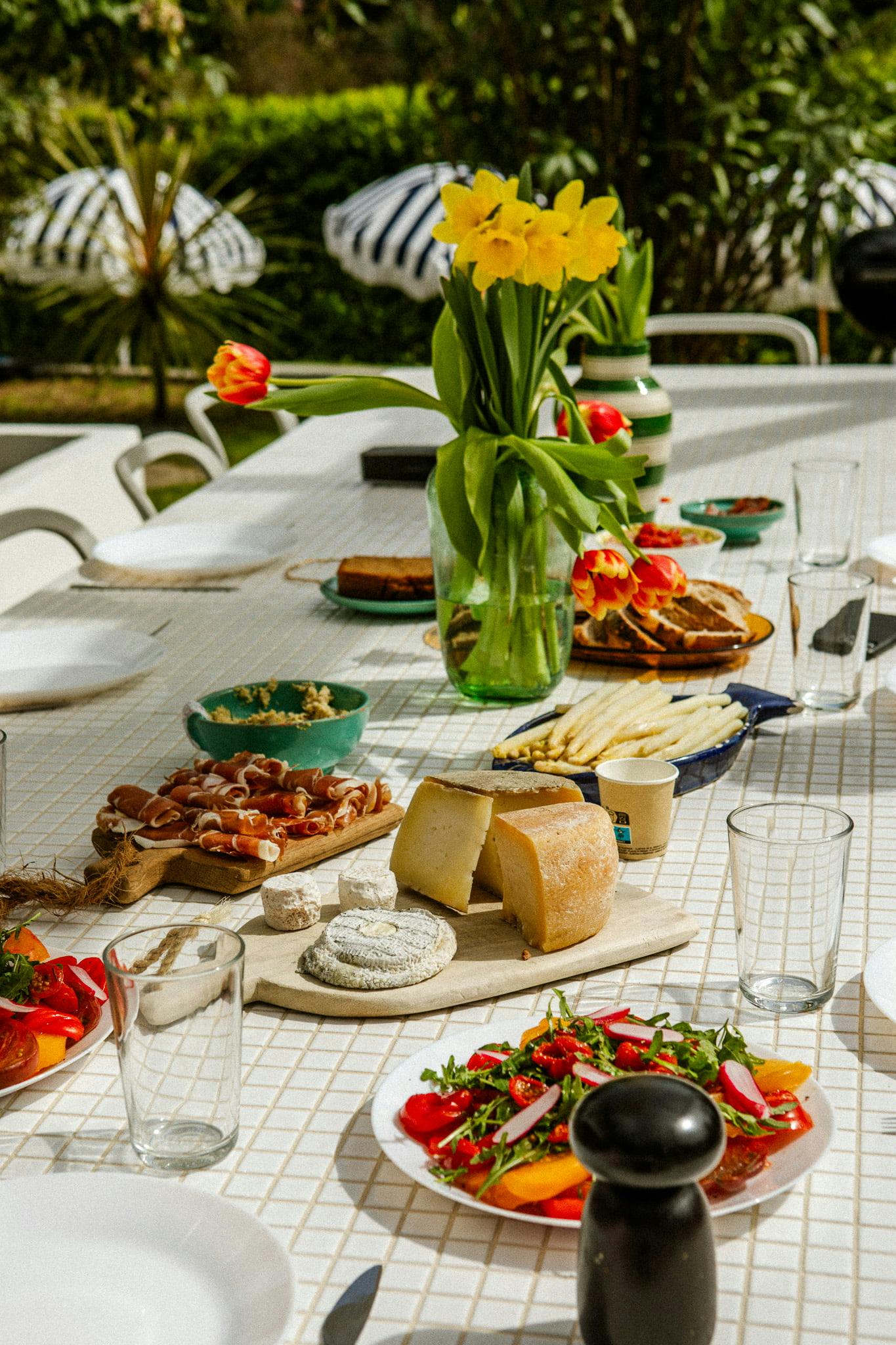 Une table à manger extérieure ensoleillée avec diverses entrées, notamment du fromage, du prosciutto, de la salade et du pain. Un vase Domino Un aux tulipes jaunes et rouges est centré sur la table. L’arrière-plan montre une verdure luxuriante et des parapluies rayés. Des assiettes, des verres et un moulin à poivre sont également sur la table.