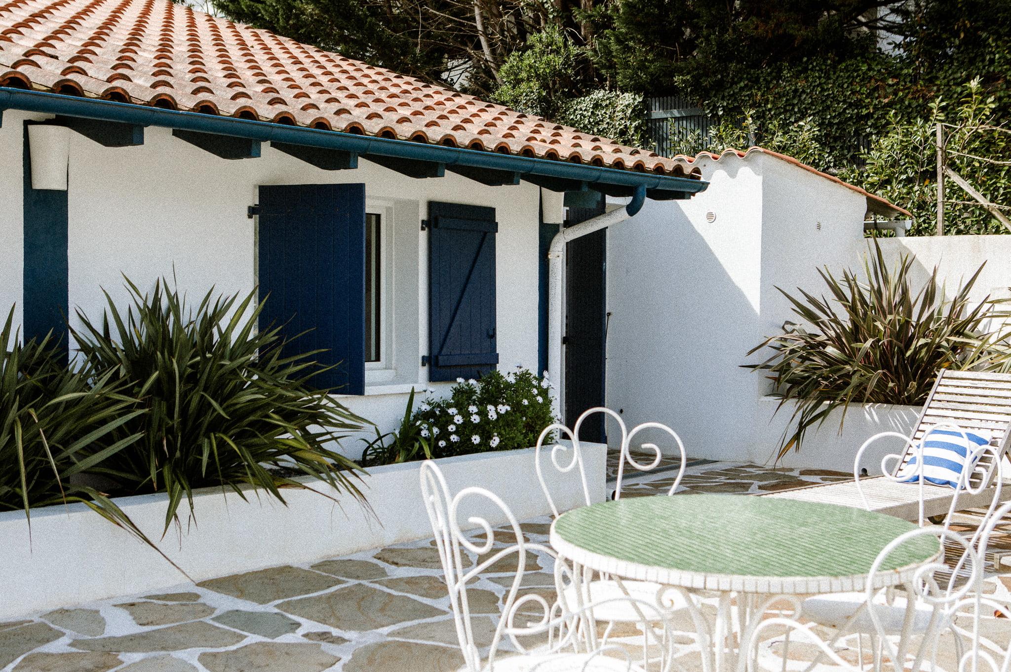 A charming white house with blue shutters and a terracotta tiled roof. The outdoor space features a stone patio, a white metal table with chairs, and lush greenery that includes spiky plants and flowering shrubs. A striped lounge chair is also visible, enhancing the relaxing ambiance of the Domino Uno style.