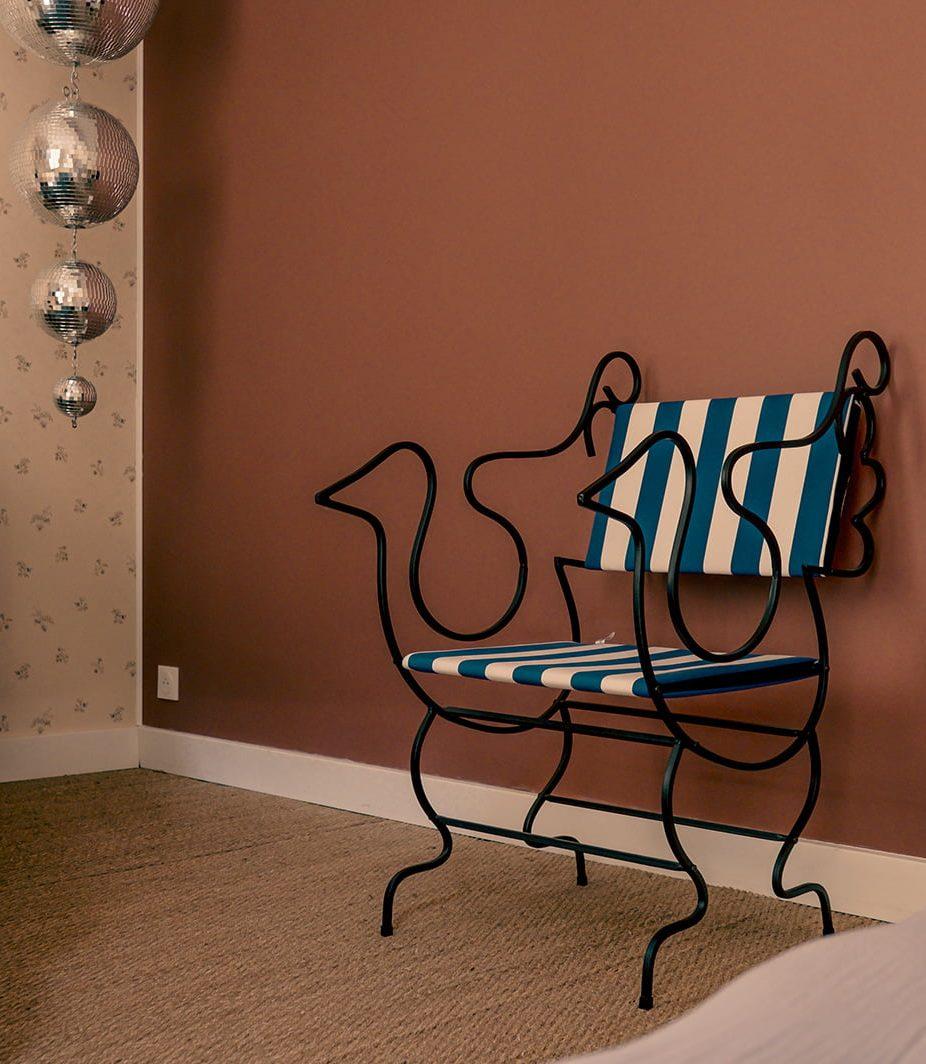 Une chaise au design unique, avec une structure en métal noir en forme de coq et des coussins rayés bleus et blancs, se dresse contre un mur brun-rosé. Trois boules disco argentées sont suspendues de manière décorative dans un coin et un tapis beige recouvre le sol.