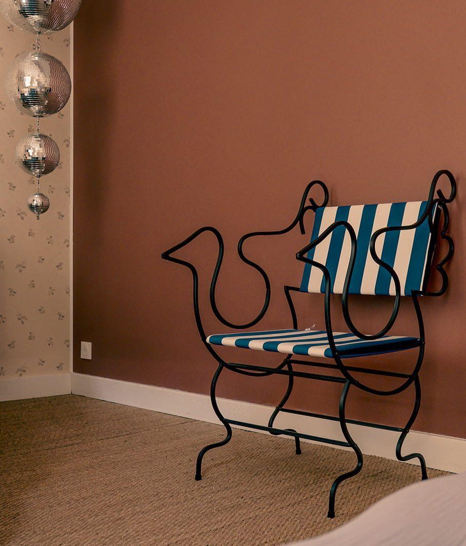 A uniquely designed chair, with a black metal frame shaped like a rooster and blue-and-white striped cushions, stands against a pinkish-brown wall. Three silver disco balls are decoratively suspended in one corner, and a beige rug covers the floor.