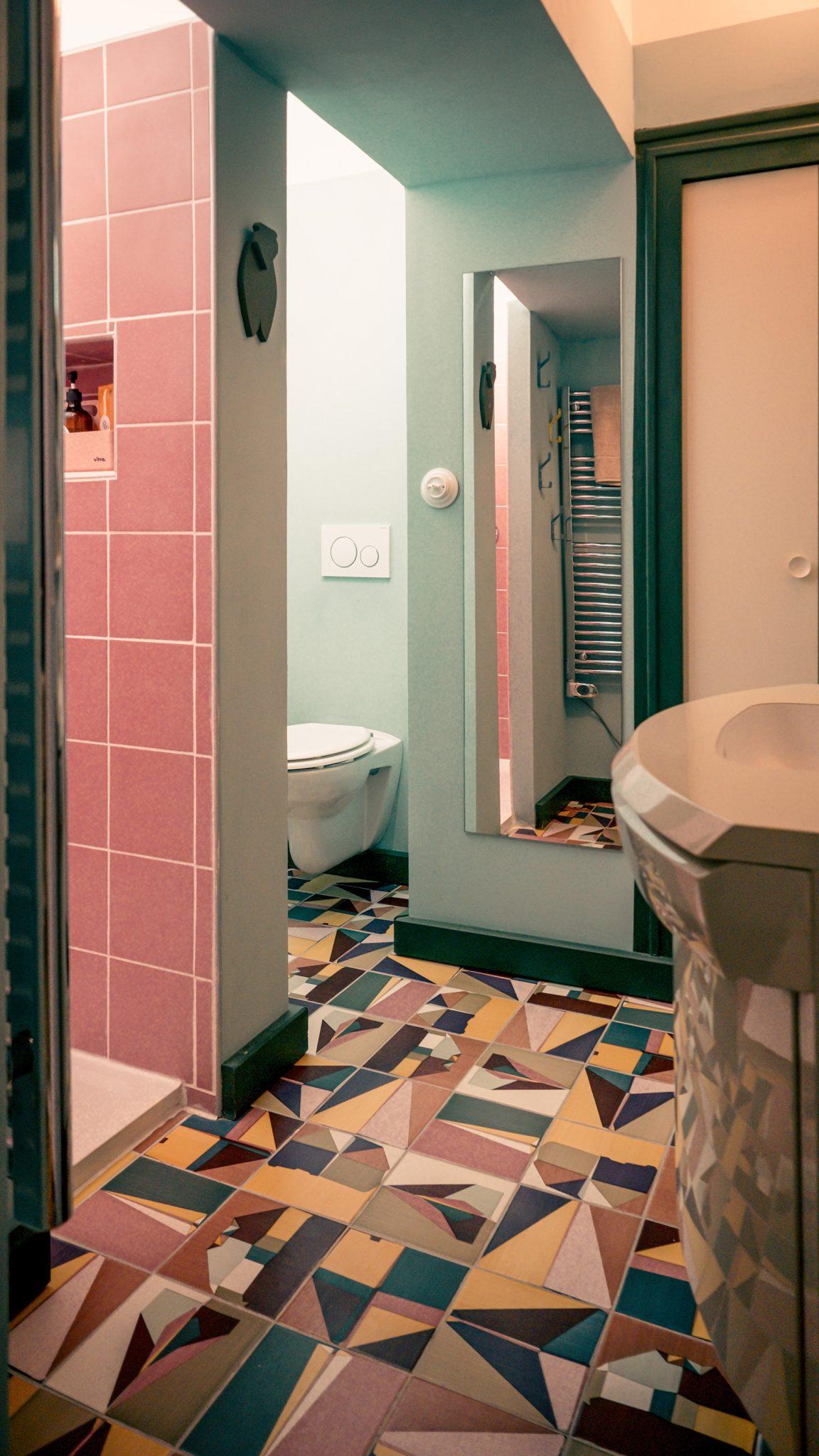 Une salle de bain avec un carrelage au sol à motifs géométriques, des toilettes blanches et un miroir mural. Les murs sont en mosaïque bleu clair et rose ornent la zone de douche. Il y a un lavabo blanc avec un plan de travail incurvé à droite et un sèche-serviettes en arrière-plan.