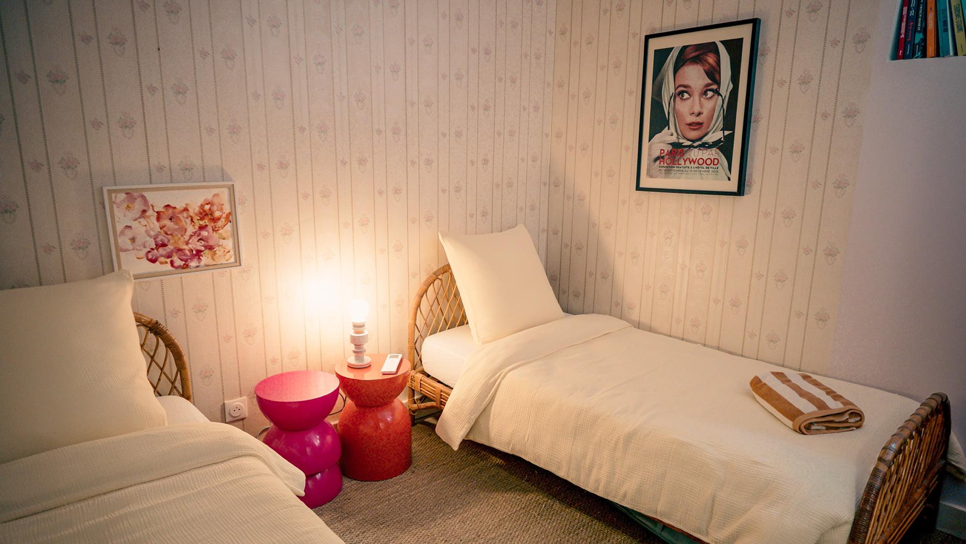 A cozy bedroom with two single beds featuring wicker frames and white bedding. Between the beds is a small round table with a pink lamp, a telephone, and a folded towel. The walls are adorned with floral wallpaper and two framed paintings, one depicting a woman and the other with flowers.