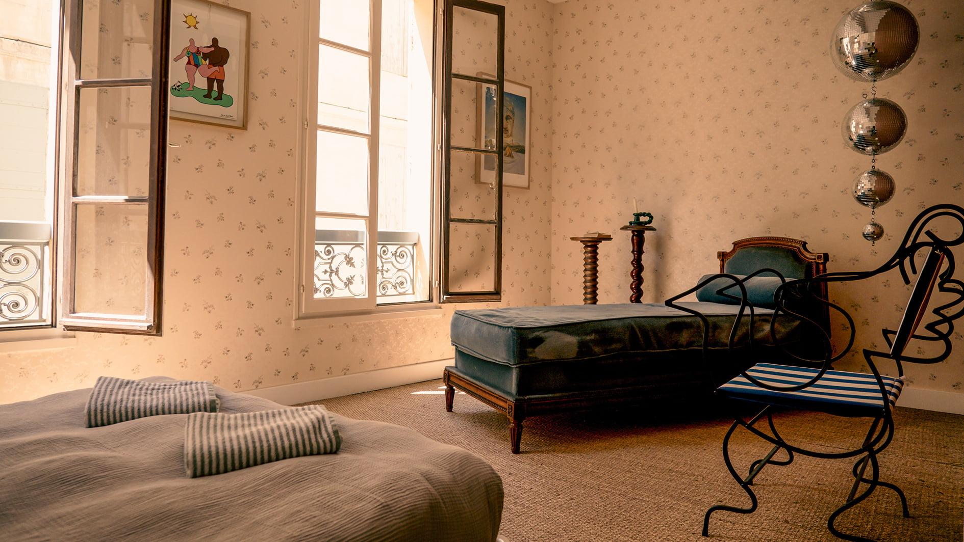 Une chambre confortable avec de grandes fenêtres, décorée de papier peint fleuri. Elle comprend un lit avec deux oreillers rayés, un deuxième lit avec un revêtement bleu sarcelle, une élégante chaise noire, des boules argentées flottantes et des œuvres d'art encadrées sur les murs, créant un mélange d'esthétique moderne et classique.