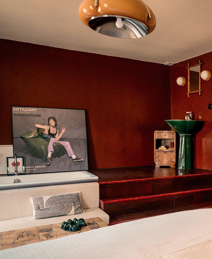 A cozy room with brown walls features a large poster of a woman partially leaning against the wall. A green sink is installed in one corner, with a small mirror hanging above it. The room is decorated with modern furniture and a brown ceiling light.