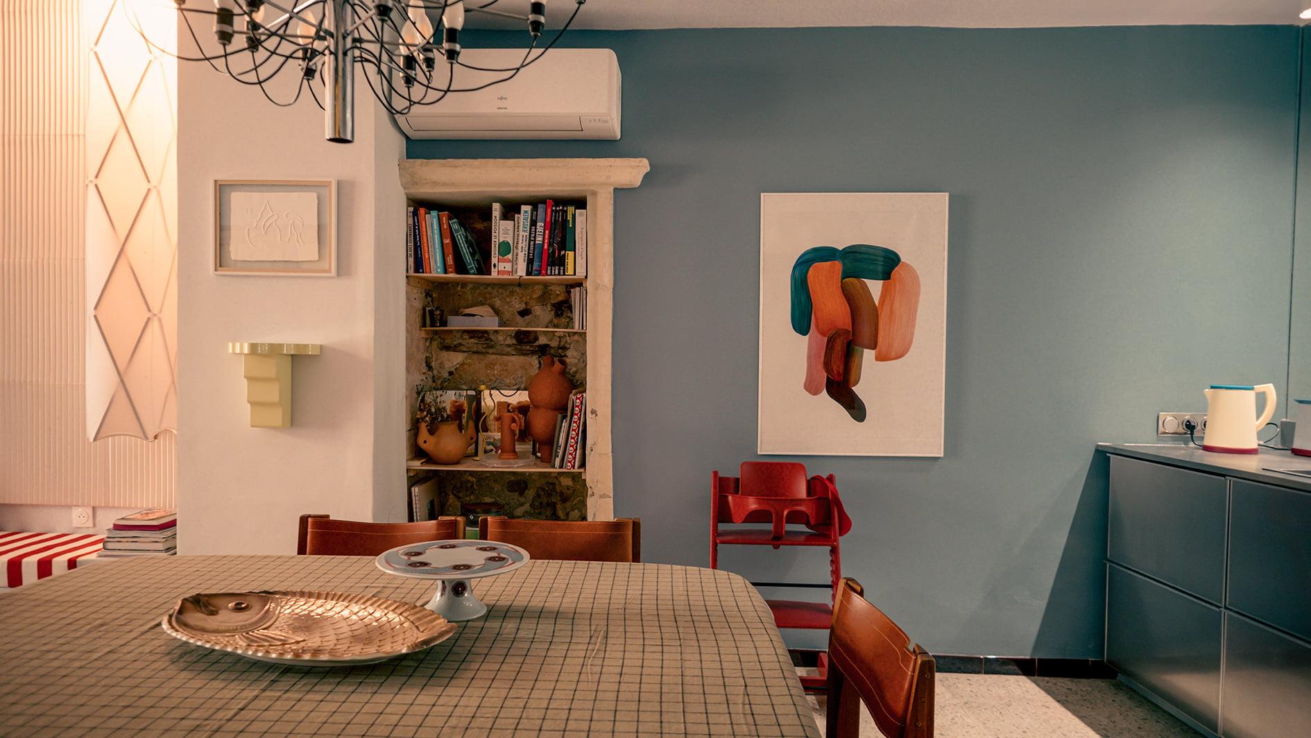 Salle à manger confortable avec une table en bois et des chaises rouges. Un lustre moderne est suspendu au plafond. La pièce présente un mur d'accent bleu avec des œuvres d'art abstraites. Il y a une étagère intégrée avec des livres et des objets décoratifs, et un climatiseur au-dessus.