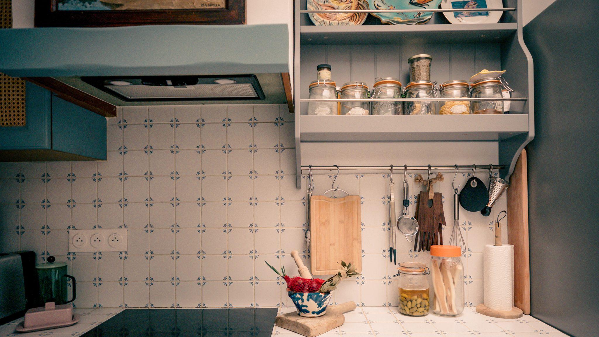 A cozy kitchen nook with a white tiled backsplash, open shelves holding spice jars and ingredients, hanging utensils, a wooden cutting board, and fresh produce in a blue bowl on the counter. An electric stove is on the left side.