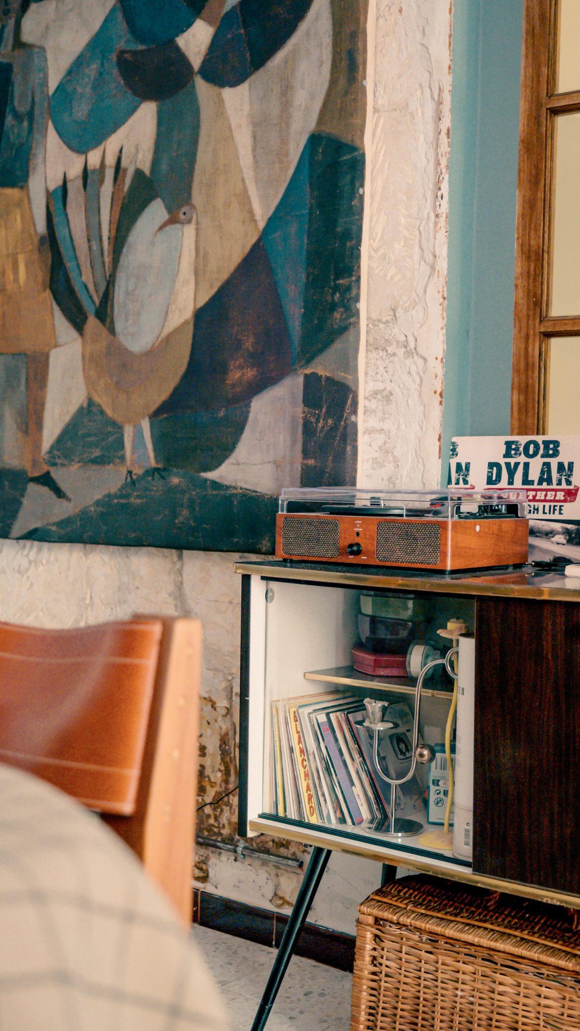 A cozy room features a mid-century modern cabinet holding a record player and vinyl records. Above it, a large abstract painting is hung on the wall. A wicker basket is placed underneath the cabinet, and part of a wooden chair is visible in the foreground.