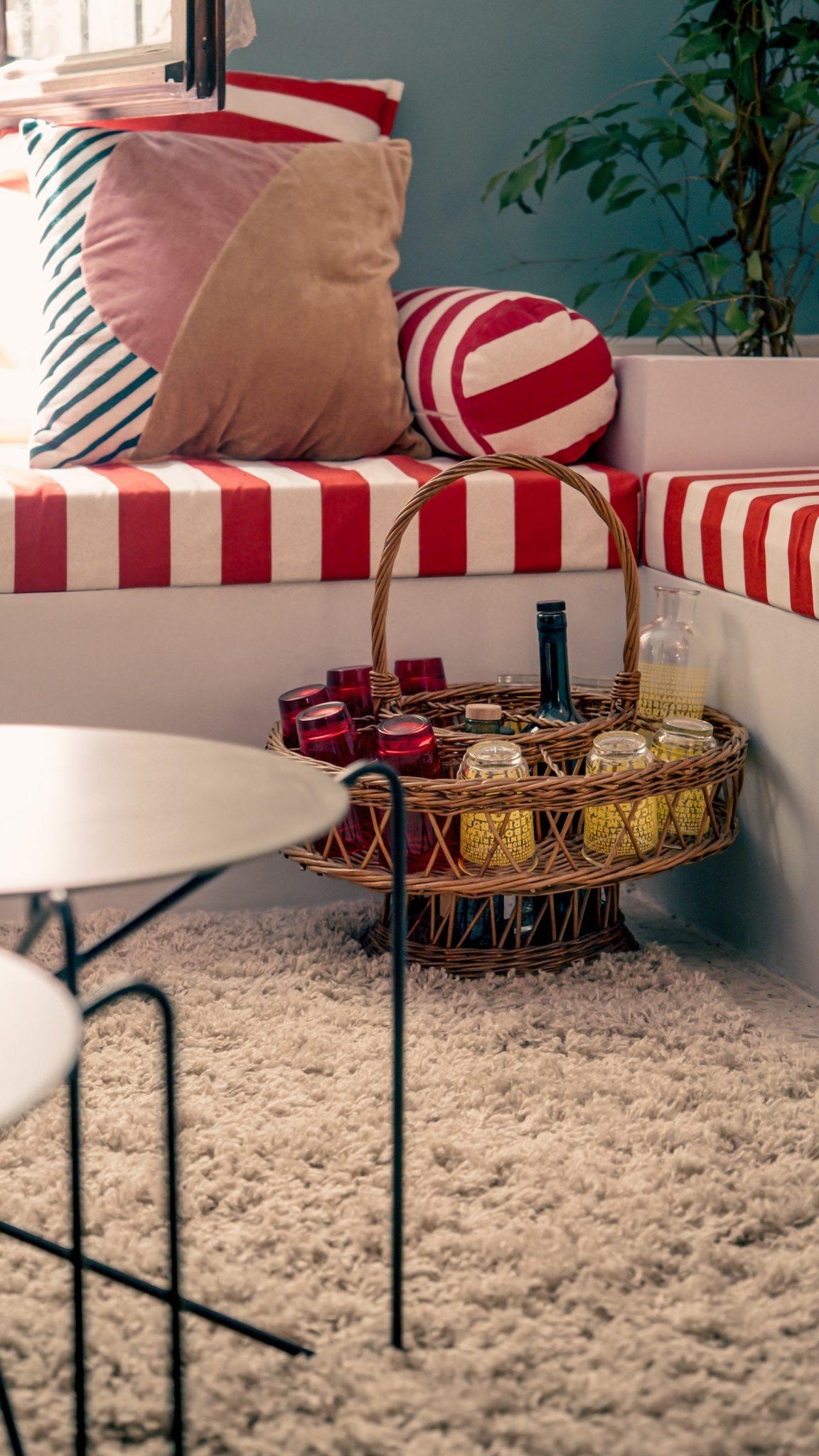 A cozy nook with a cushioned seating area upholstered in red and white stripes, adorned with various pillows. A wicker basket filled with bottles and lemons sits on a soft, light-colored rug. A small minimalist table is in the foreground, and a plant is visible in the background.