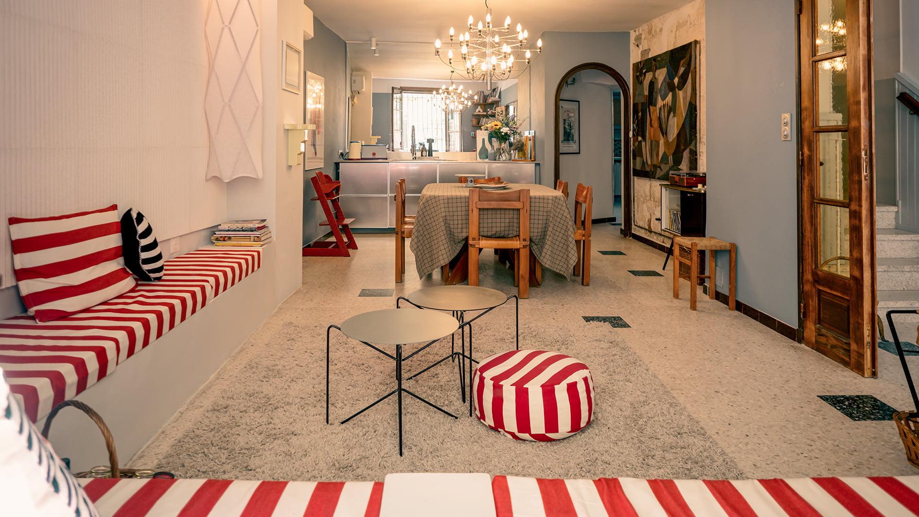 A comfortable and well-lit living and dining room with cushions and décor in red and white stripes. The space includes a dining table with a checkered tablecloth, a bench, a chandelier, modern furniture, and a kitchen in the background. The floor is tiled in a light color.