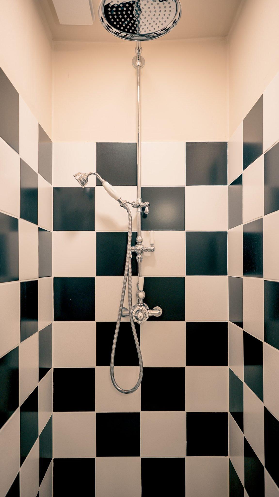 Une cabine de douche avec des carreaux à carreaux noirs et blancs sur les murs. La douche est équipée d'une grande pomme de douche ronde et d'une douchette à main montée sur une barre verticale avec des boutons de commande métalliques complexes.