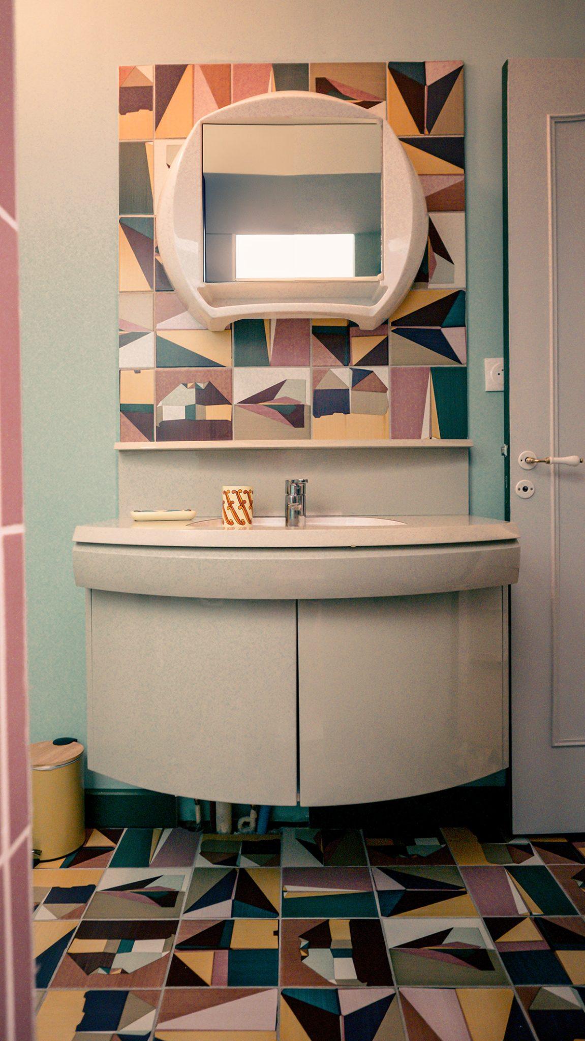 A modern-themed bathroom with geometric patterns. The mirror is circular, framed by colorful tiles in various shapes and shades. Below is a white sink with a curved cabinet. The floor mirrors the tile pattern of the mirror, continuing the geometric design throughout the space.