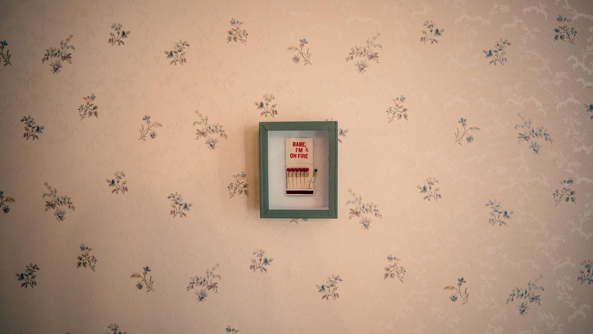 A framed cross-stitch artwork hangs on floral-patterned wallpaper. The frame is green, and the cross-stitch reads "BABE, I'M ON FIRE" with four red and white matches aligned beneath the text. The overall decor has a vintage touch.