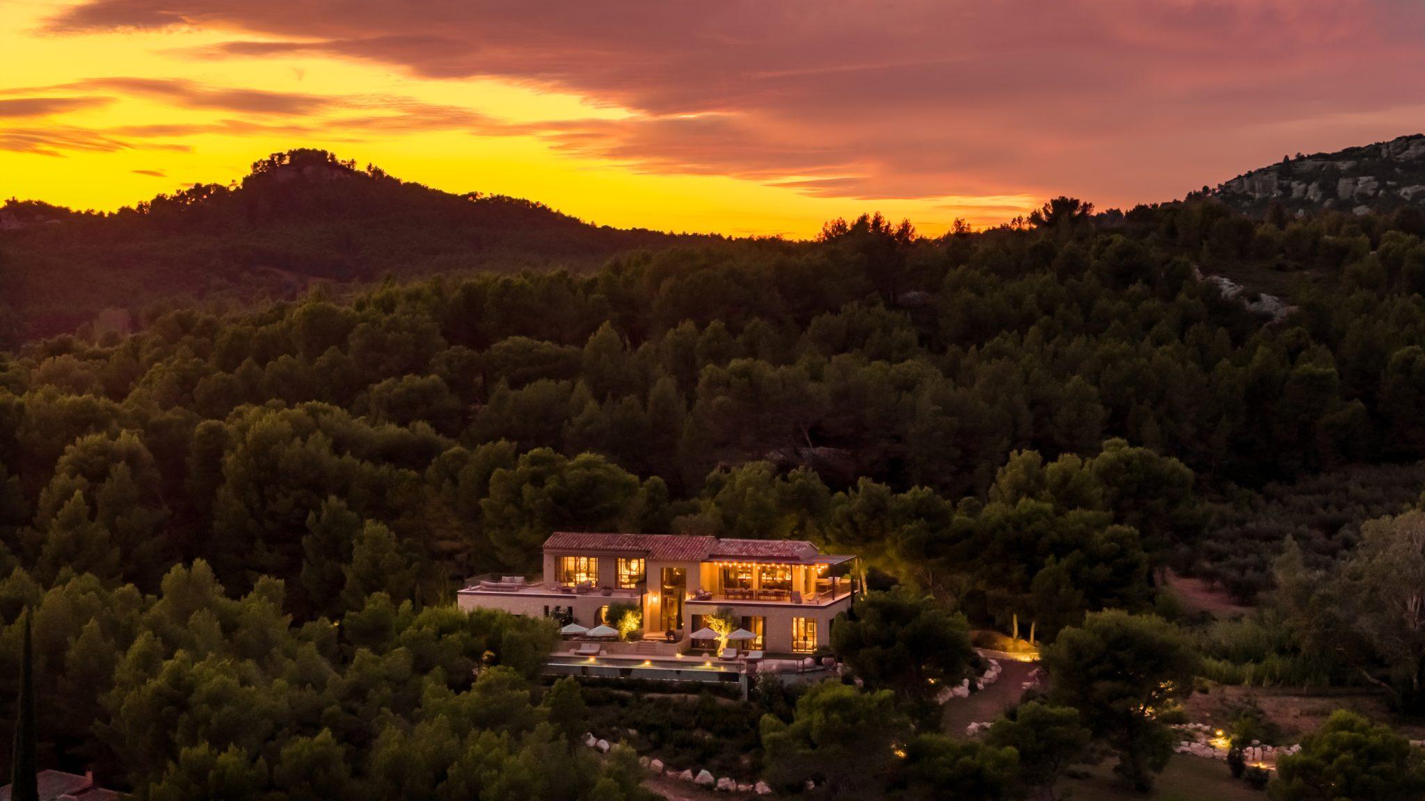 A luxurious villa with illuminated windows nestled in a lush, verdant forest is captured at sunset. The sky is bathed in orange and pink hues, creating a warm and serene atmosphere. Hills and additional wooded areas are visible in the background.