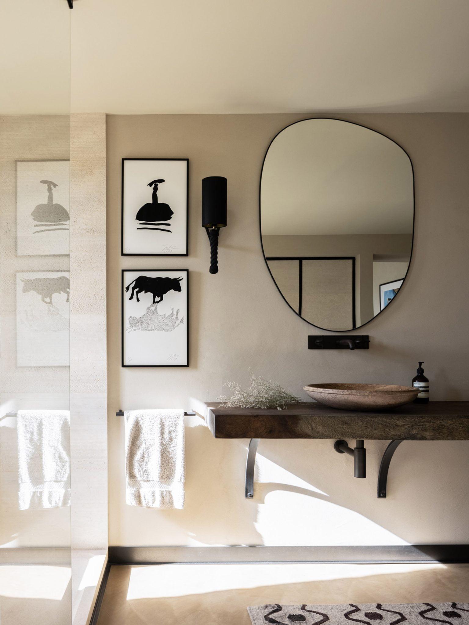 A minimalist bathroom features a wooden vanity with a stone sink, an oval mirror, and a black wall sconce. Artwork depicting abstract animal figures is hung on the wall. A hand towel is draped over a holder next to a glass shower enclosure.