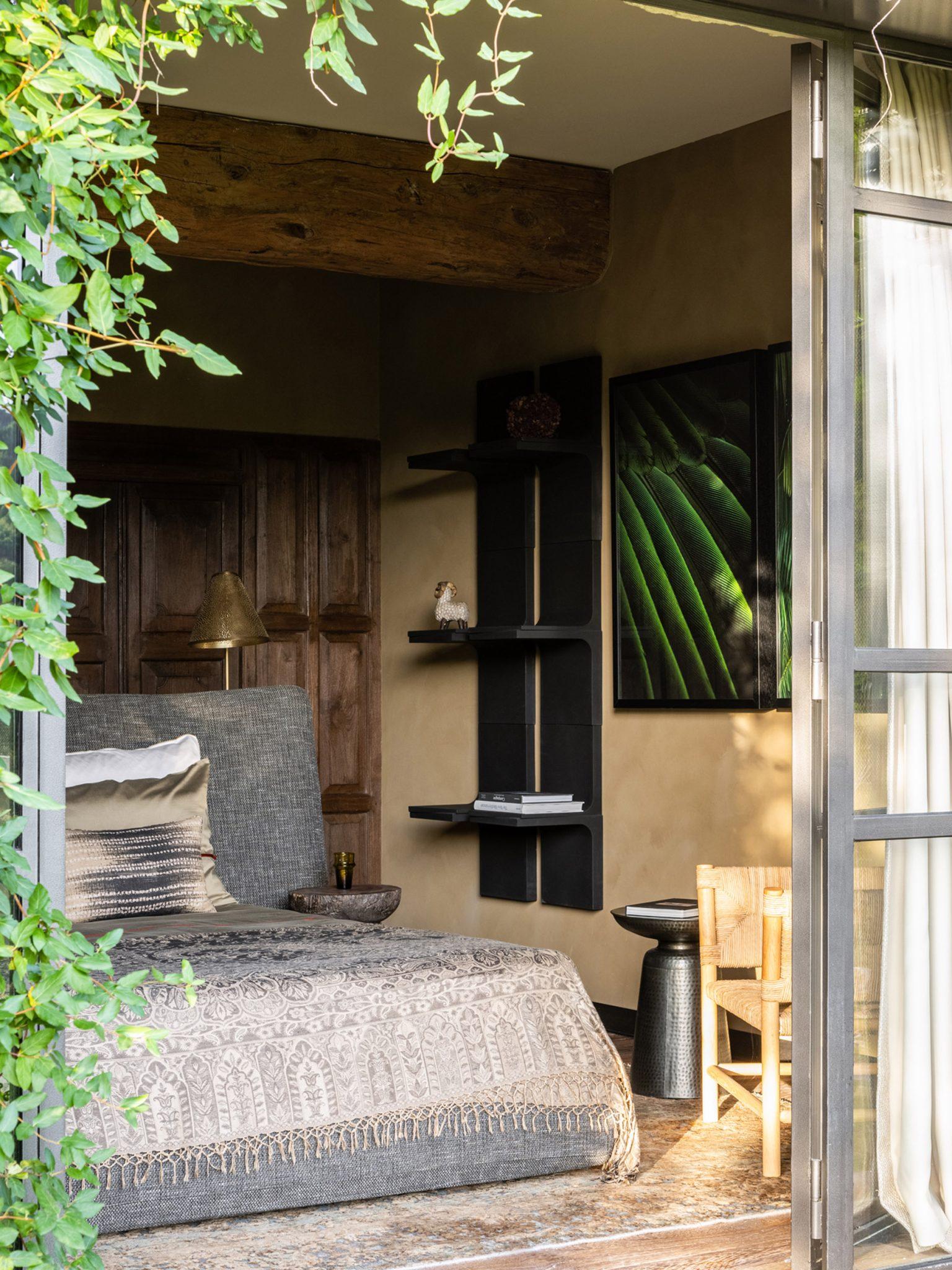 Une chambre confortable aux tons terreux comprenant un lit rembourré avec une literie à motifs, une tête de lit en bois, des étagères murales, des œuvres d'art encadrées de feuilles vertes, une chaise en bois, une table d'appoint et de la verdure visible à travers une porte vitrée ouverte.