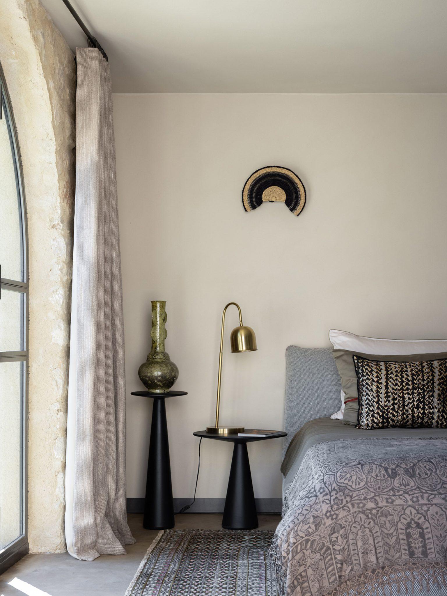 A cozy bedroom features a bed with patterned bedding and matching pillows. Beside the bed are two black nightstands, each with a green vase, a brass desk lamp, and a decorative wall piece above. Sunlight filters through a large arched window with beige curtains.