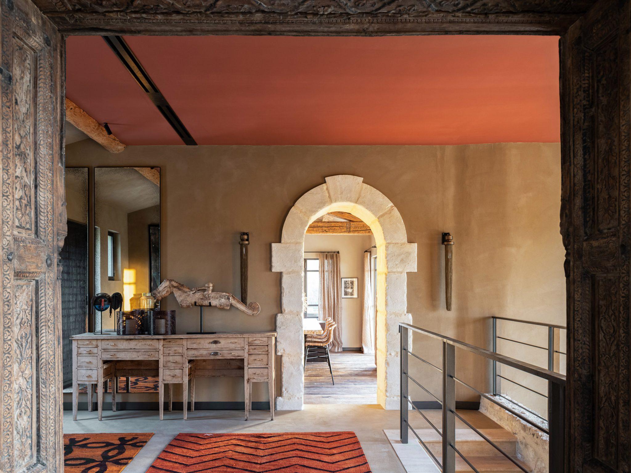 A warm and rustic interior with a wooden desk and a mirror against a textured wall. An arched doorway with a light stone frame leads to a cozy room beyond. A patterned red rug is laid on the floor, and a metal railing lines the adjacent staircase, under a terracotta-colored ceiling.