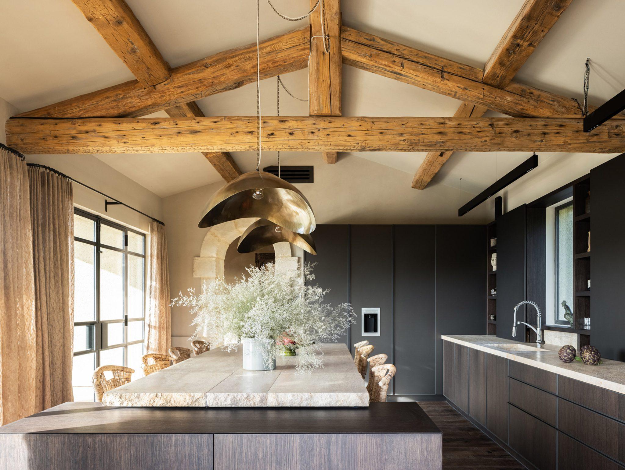 A modern kitchen with dark cabinetry, a large central island with a marble countertop and sink. The room features exposed ceiling beams and large pendant lights. A dining table with wicker chairs and decorative plants is situated next to a set of windows with curtains.