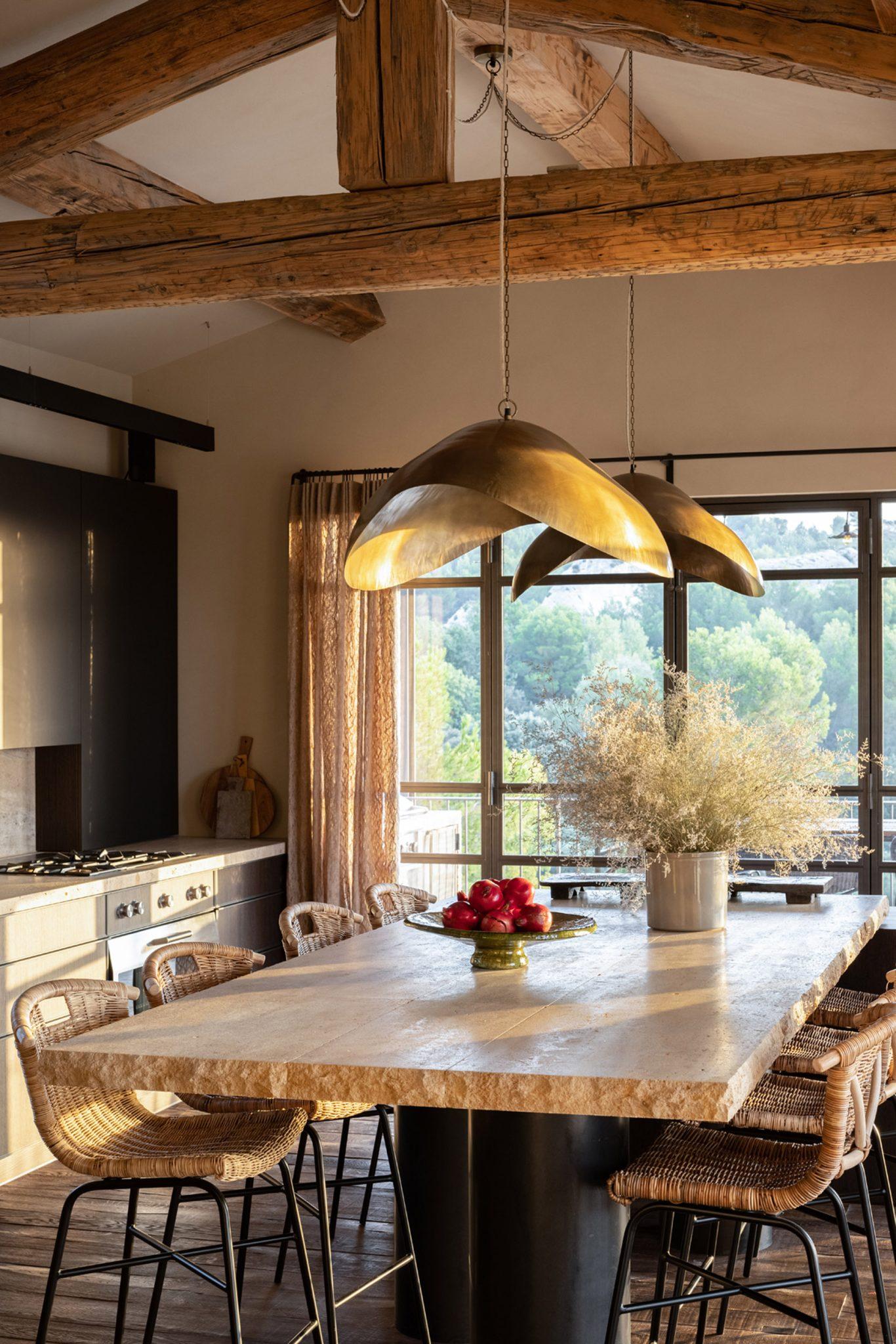 A rustic kitchen featuring a large wooden island with wicker bar stools and two large brass pendant lights. The island is adorned with a bowl of red apples and a vase of dried plants. The space has wooden beams on the ceiling and a window offering a panoramic view of the outdoors.