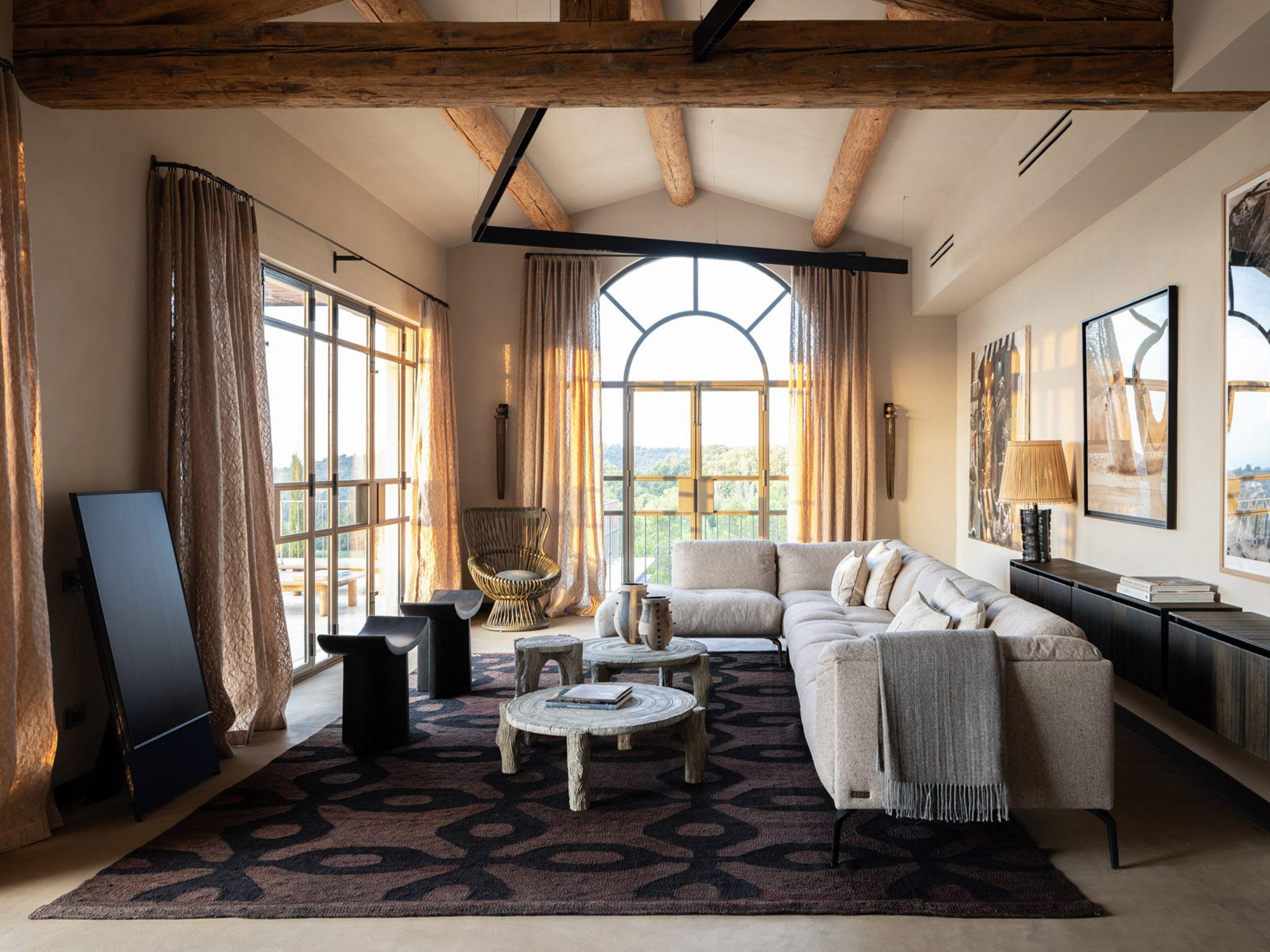 An elegant living room with high ceilings and exposed wooden beams. The space features beige and neutral-toned decor, including a plush sectional sofa, a coffee table, and various modern furniture pieces. Floor-to-ceiling windows allow natural light to flood the room.