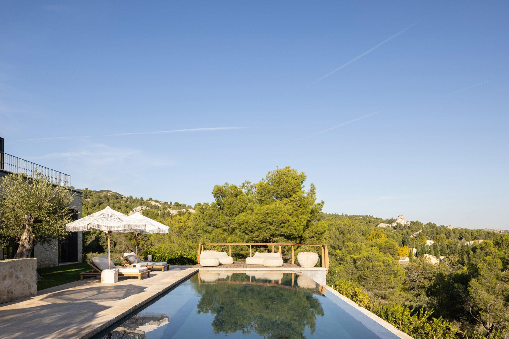 A serene outdoor pool with clear blue water reflecting the sky is surrounded by greenery and trees. Along the edge of the pool, several comfortable white lounge chairs and umbrellas provide shade. In the background, rolling hills covered in trees are visible under a clear blue sky.