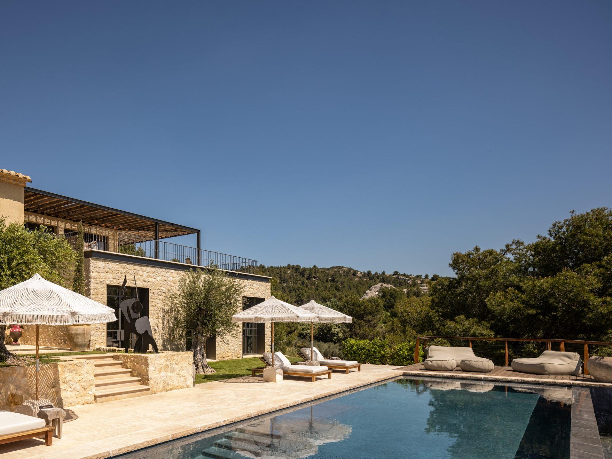 A luxurious villa with natural stone walls features a sparkling outdoor pool surrounded by lounge chairs with white umbrellas. The villa overlooks a picturesque landscape filled with lush greenery under a clear blue sky.