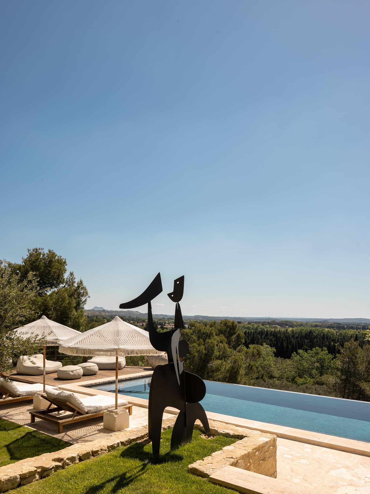 A modern outdoor pool area with white lounge chairs and umbrellas on a stone terrace. A black abstract sculpture stands in the foreground. The serene landscape features trees and a distant view of rolling hills under a clear blue sky.