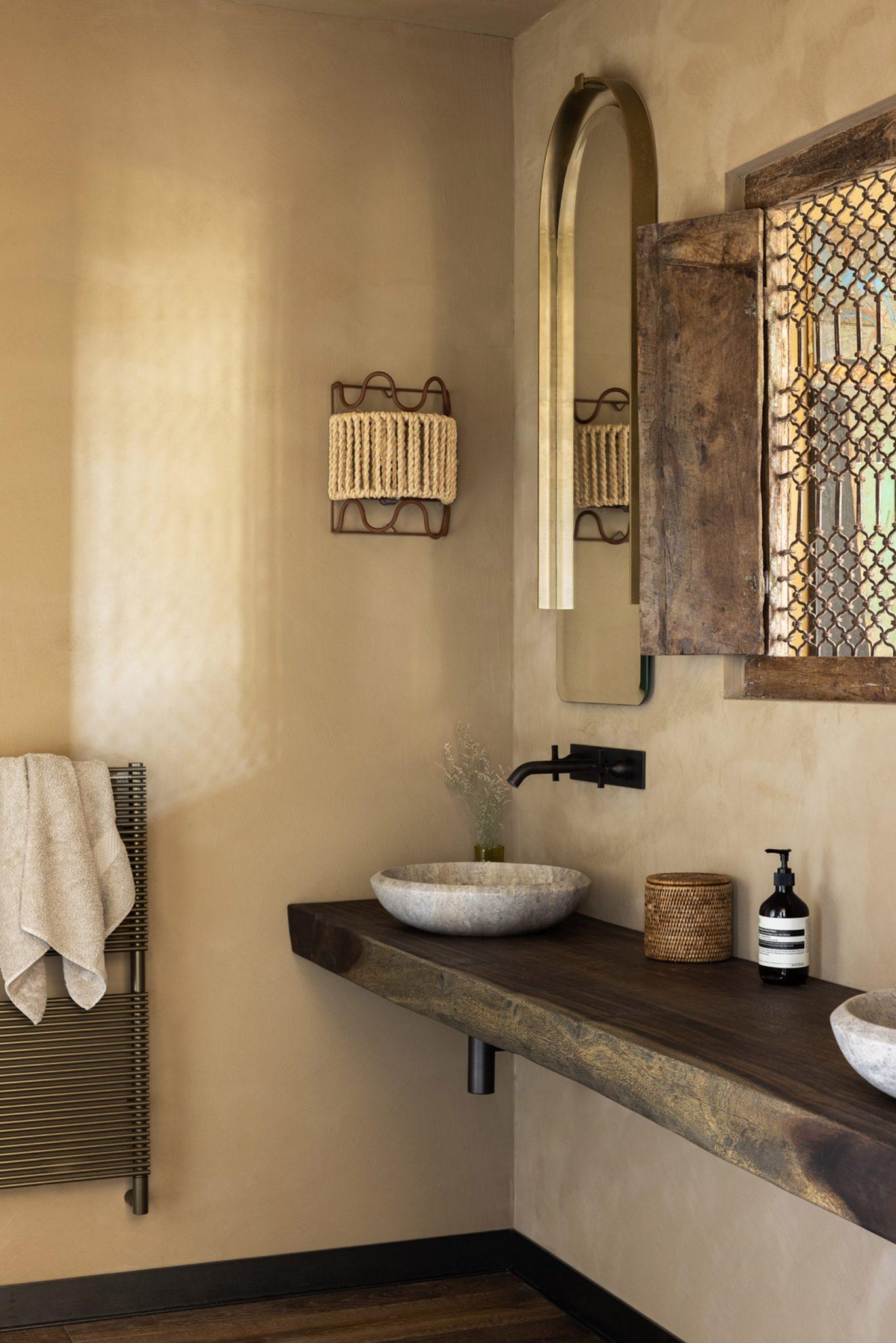 A modern bathroom with beige walls features a rustic wooden vanity with two large stone sinks and black faucets. Above the sinks is a large curved mirror. The wall sconce is covered with woven fabric, and a towel hangs from a black radiator.