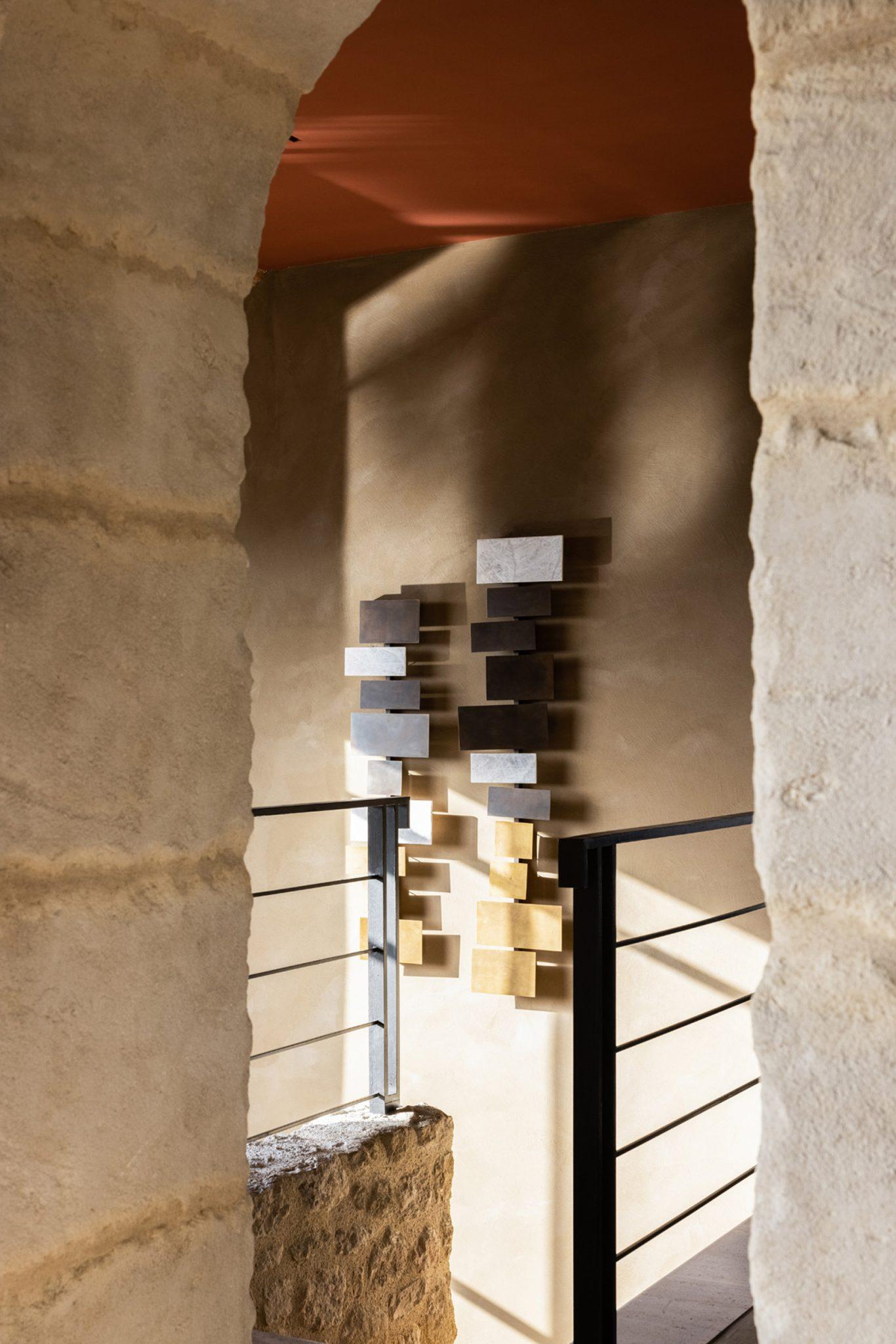 A view through an archway reveals a modern, abstract wall sculpture composed of rectangular blocks of various colors and sizes. The sculpture is mounted on a beige wall, illuminated by natural light. Iron railings and stone walls frame the composition.