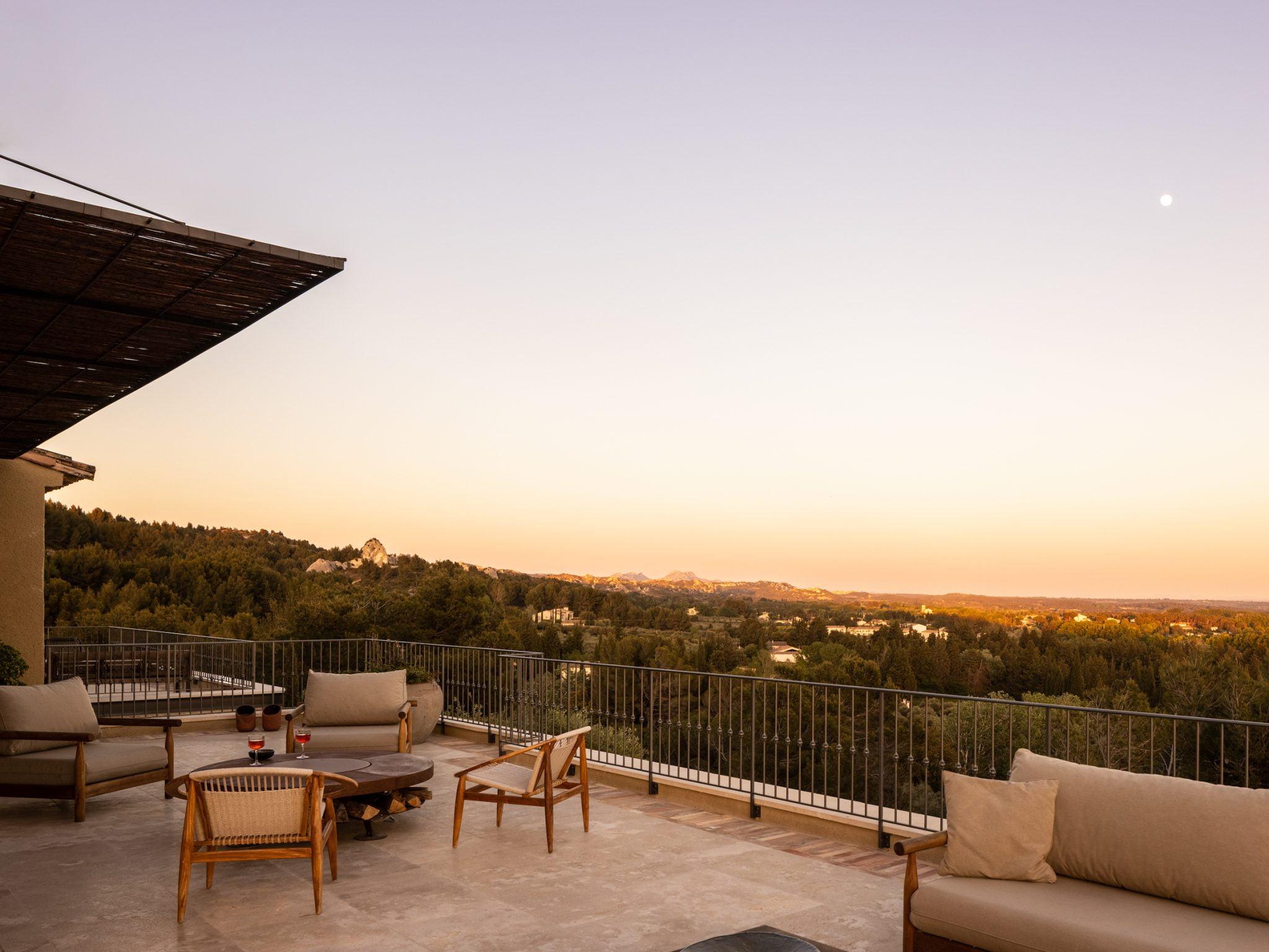 A spacious terrace with comfortable outdoor furniture, including a loveseat, armchairs, and a coffee table, overlooks a picturesque landscape of rolling hills and distant mountains at sunset. The sky is clear with a visible moon, and the area is softly illuminated by warm hues.