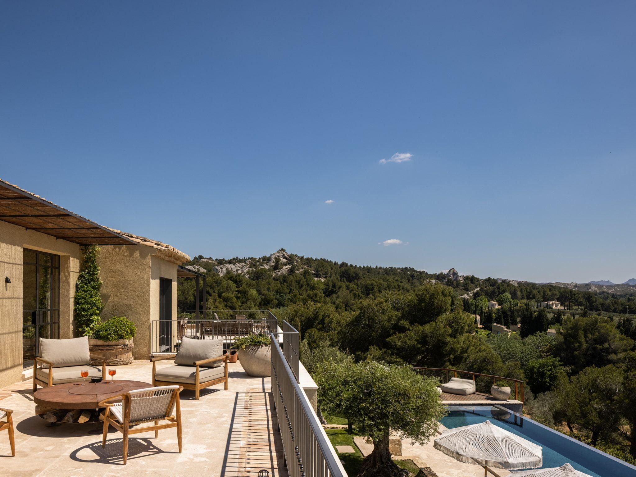 Une terrasse luxueuse avec un coin salon, comprenant des chaises rembourrées et une table en bois. Elle donne sur un vaste paysage boisé, une piscine privée et des montagnes lointaines sous un ciel bleu clair. L'atmosphère est lumineuse et tranquille.