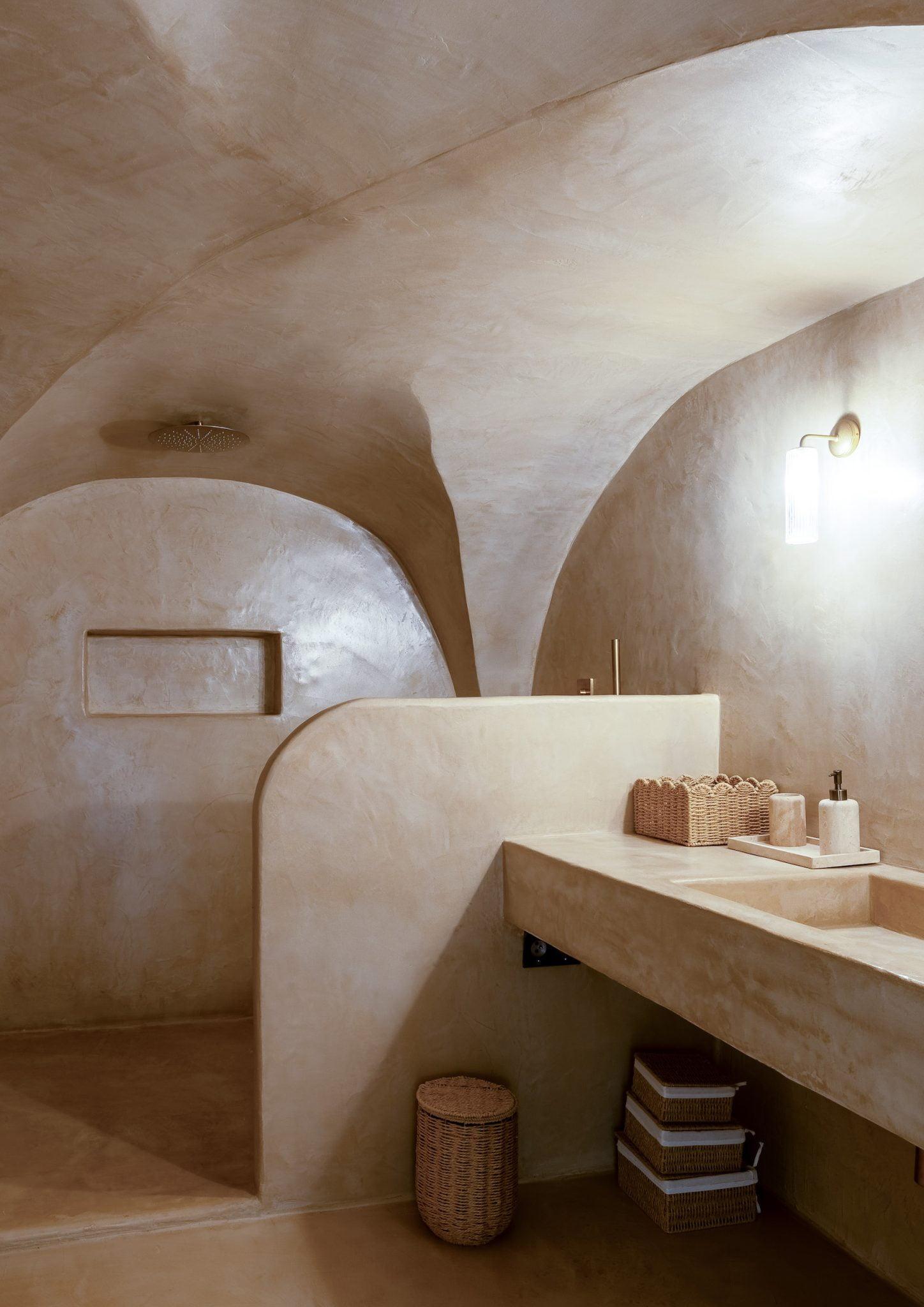 A minimalist bathroom with smooth stucco walls and ceiling in beige tones. It features a large countertop sink, wicker baskets, toiletries, and a modern backlit wall sconce. The space includes a built-in shower with a high showerhead and a recessed shelf.