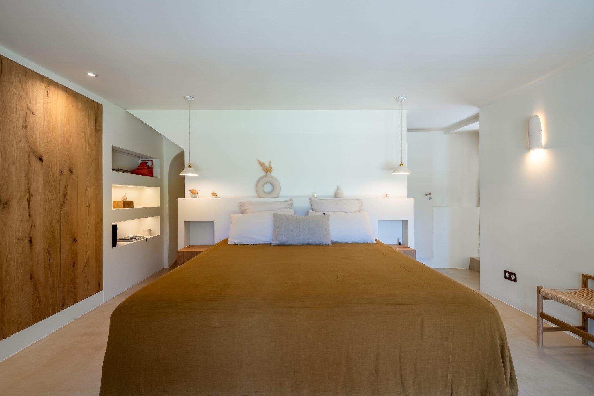 A minimalist bedroom with a large bed centered and covered in brown bedding. The room features wooden accents, built-in shelves with minimalist décor, white walls, pendant lamps, and soft lighting. A wooden wardrobe and a simple chair are also visible.