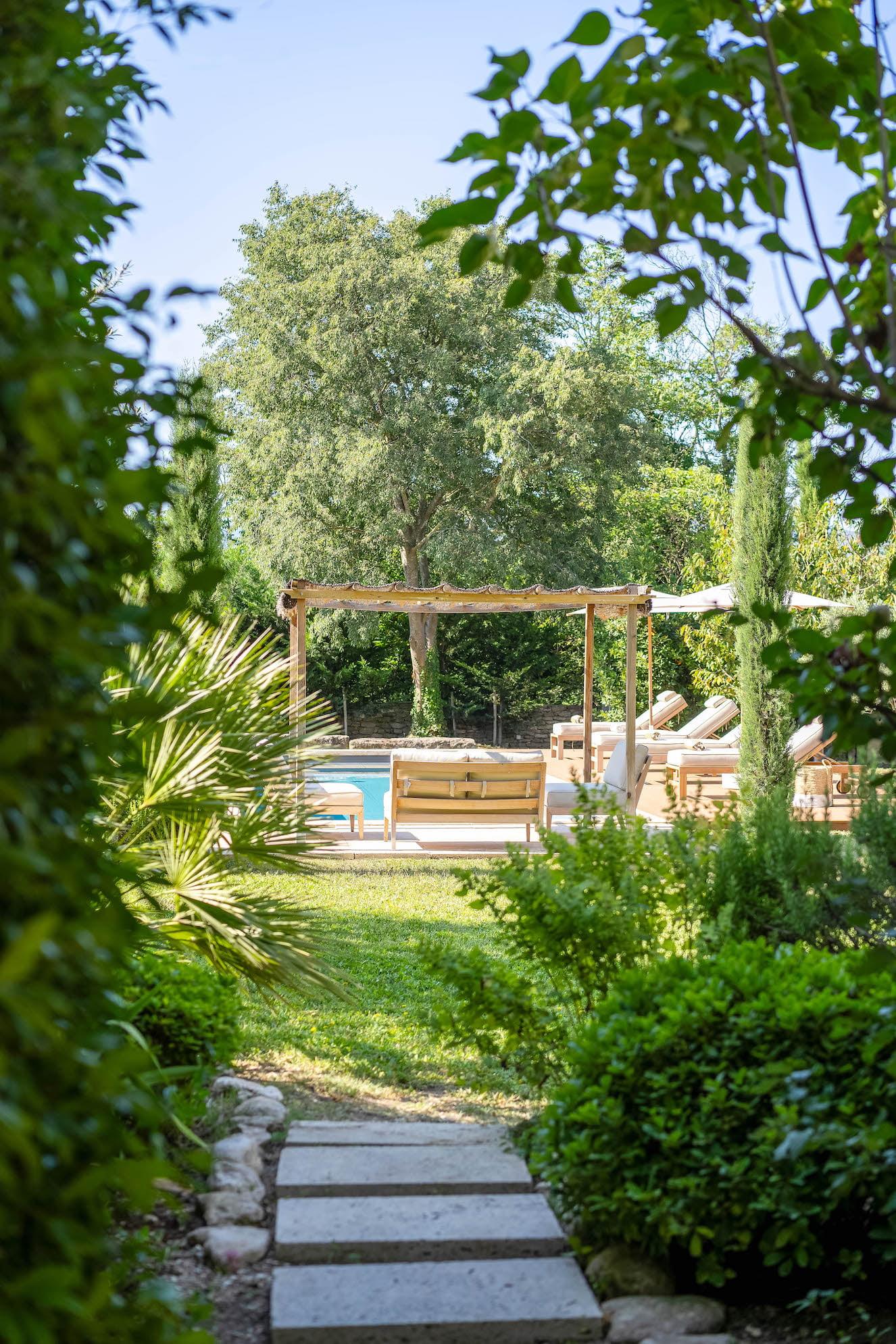 A serene garden path made of stone slabs leads to a relaxing pool area with lounge chairs under a wooden pergola. The landscape is surrounded by lush green plants and trees, creating a peaceful and nature-filled atmosphere.