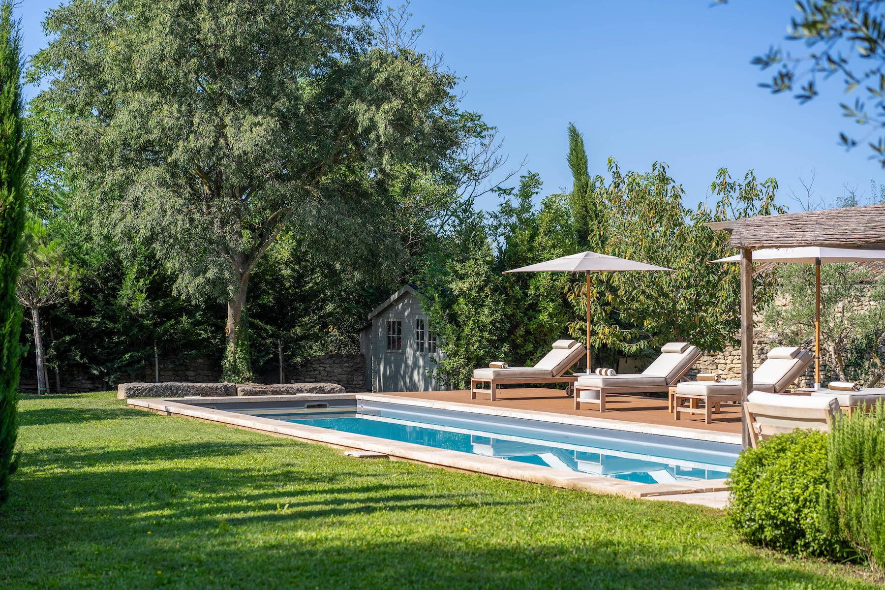 A serene outdoor pool scene features several lounge chairs with cushions under a large umbrella on a wooden deck. The pool is surrounded by lush green lawn and tall trees. A charming little cabana is nestled in the background amidst the greenery.