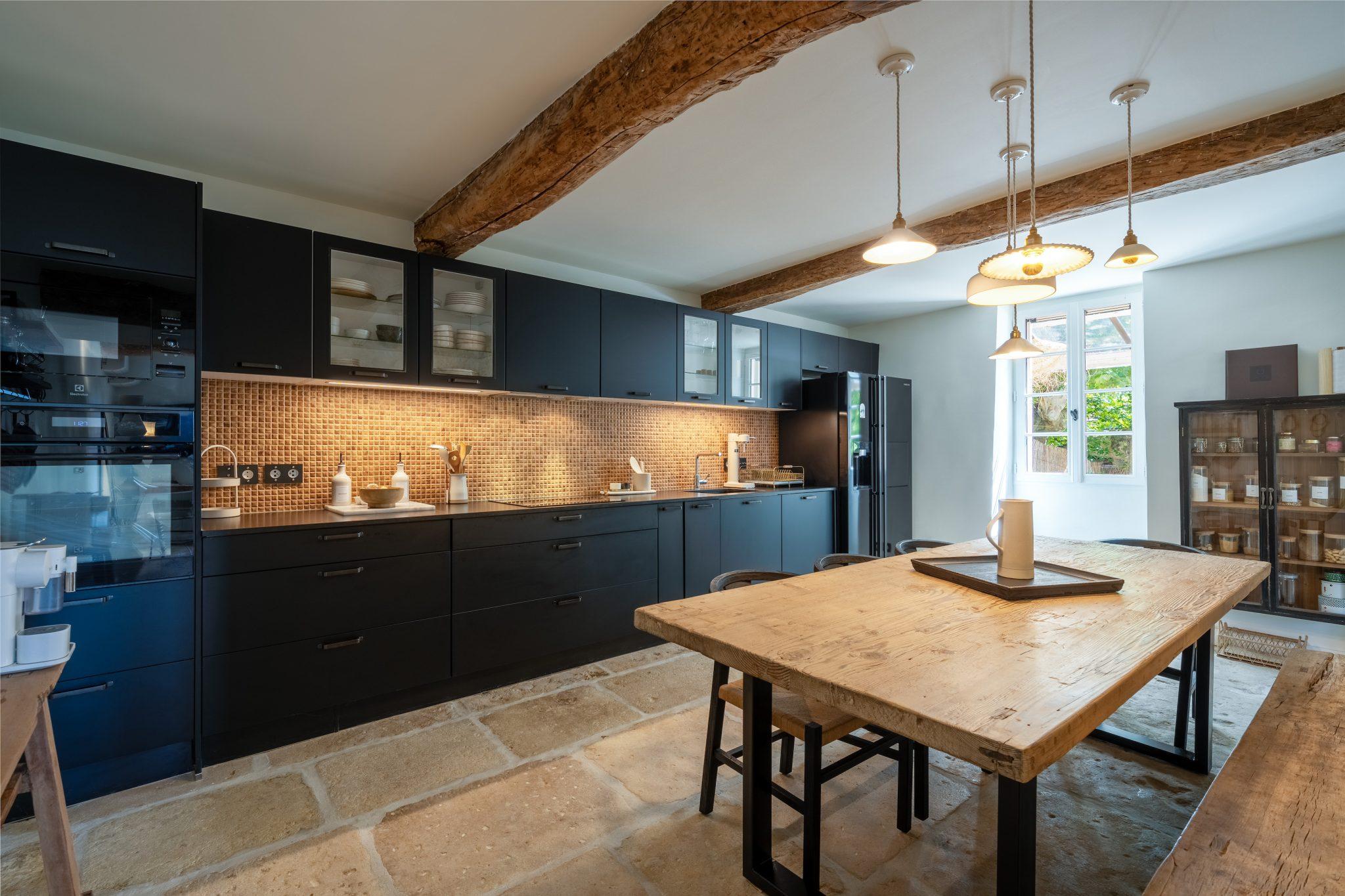 A spacious and modern kitchen with dark cabinetry, a wooden dining table, and exposed wooden beams on the ceiling. The backsplash features small beige tiles. Pendant lights hang above the table. Large windows allow natural light to flood in, and the floor is covered with large stone tiles.