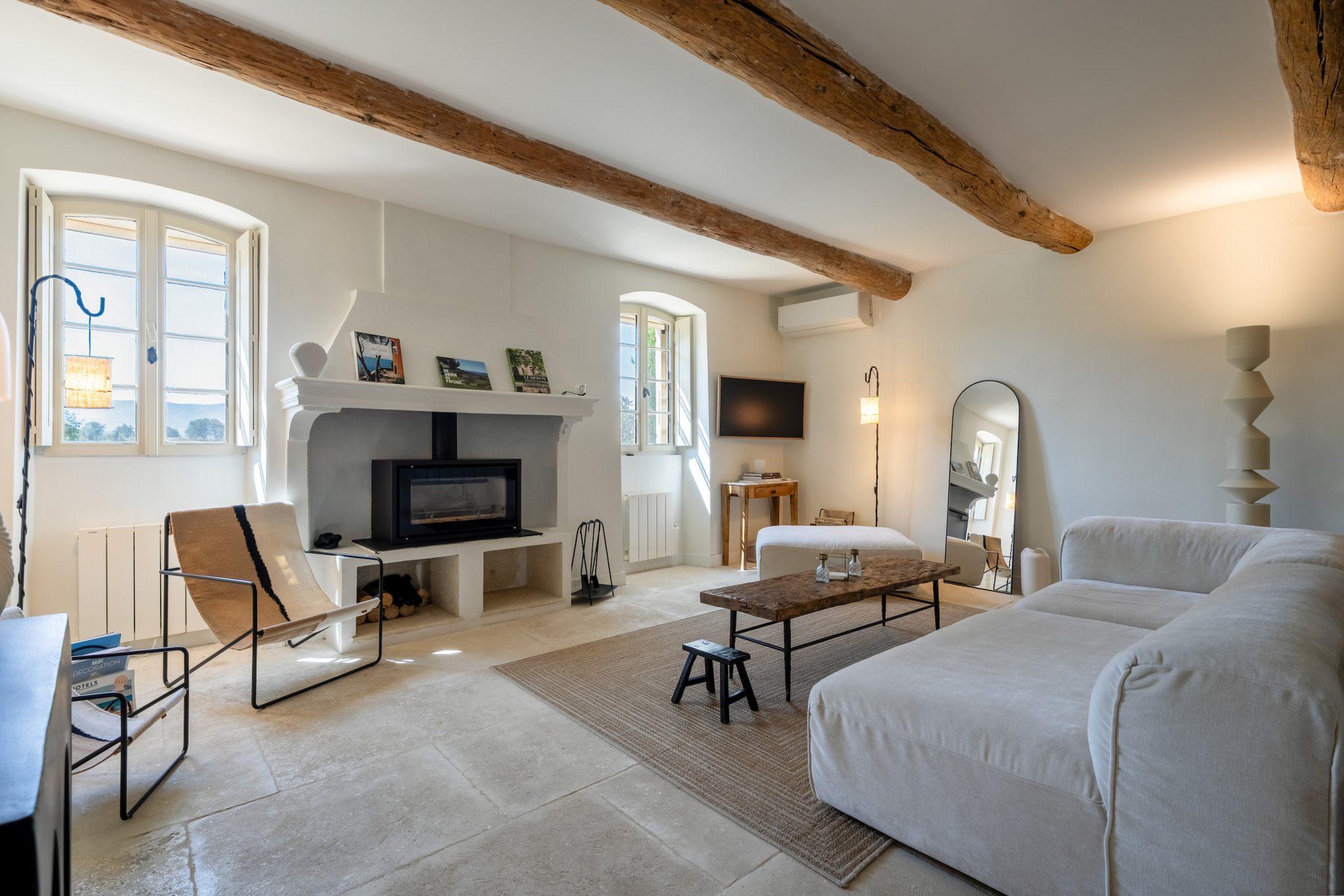 A cozy living room with rustic wooden beams on the ceiling, a large white sectional sofa, a modern fireplace, a wall-mounted flat-screen TV, a wooden coffee table, a woven chaise lounge, and a large mirror leaning against the wall. Natural light streams in through two windows.