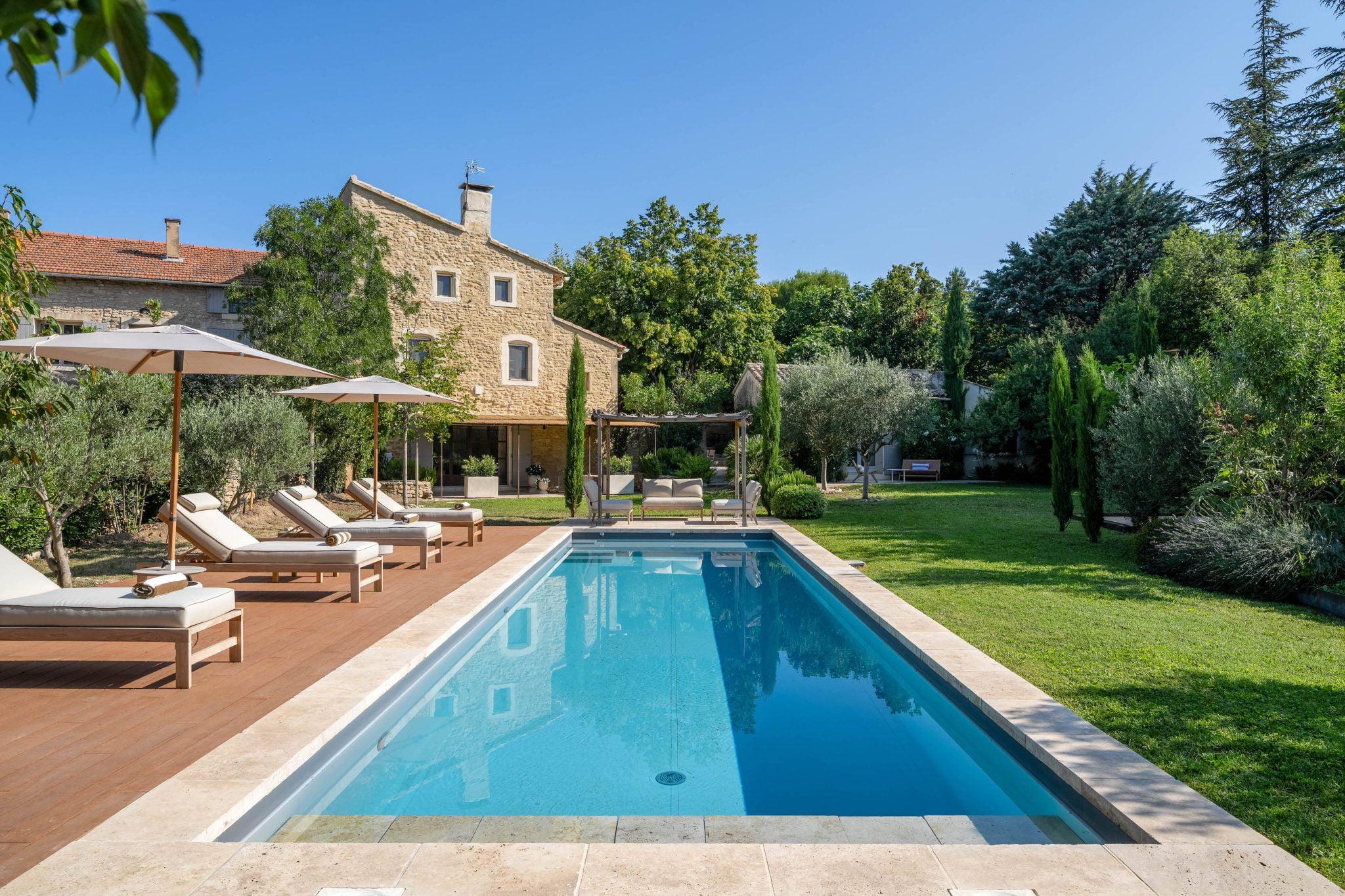 A serene backyard includes a rectangular pool surrounded by lounge chairs with umbrellas. The pool is framed by a wooden deck and lush green lawn. A stone house is visible in the background, along with tall trees and shrubs. The sky is clear and blue.