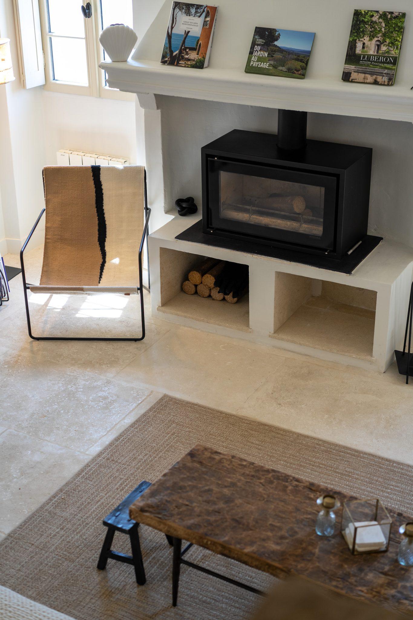 Un coin salon avec un poêle noir incrusté dans le mur blanc, un fauteuil en rotin et des cadres sur une étagère murale.