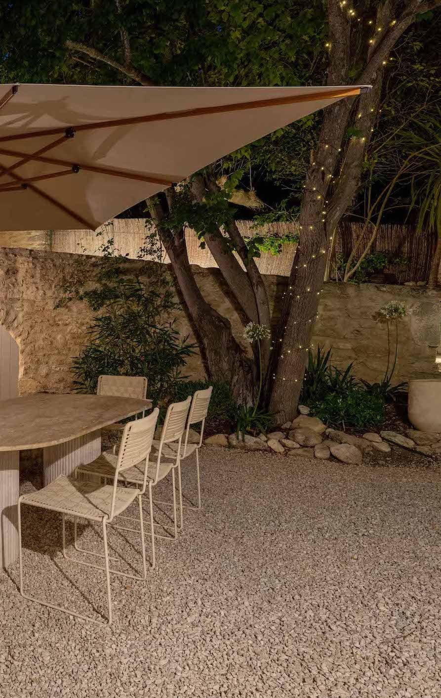 A peaceful outdoor patio with a large umbrella shading a stone table and white chairs. The space is illuminated by string lights wrapped around a tree, creating a warm ambiance. The décor includes a stone wall and lush greenery.