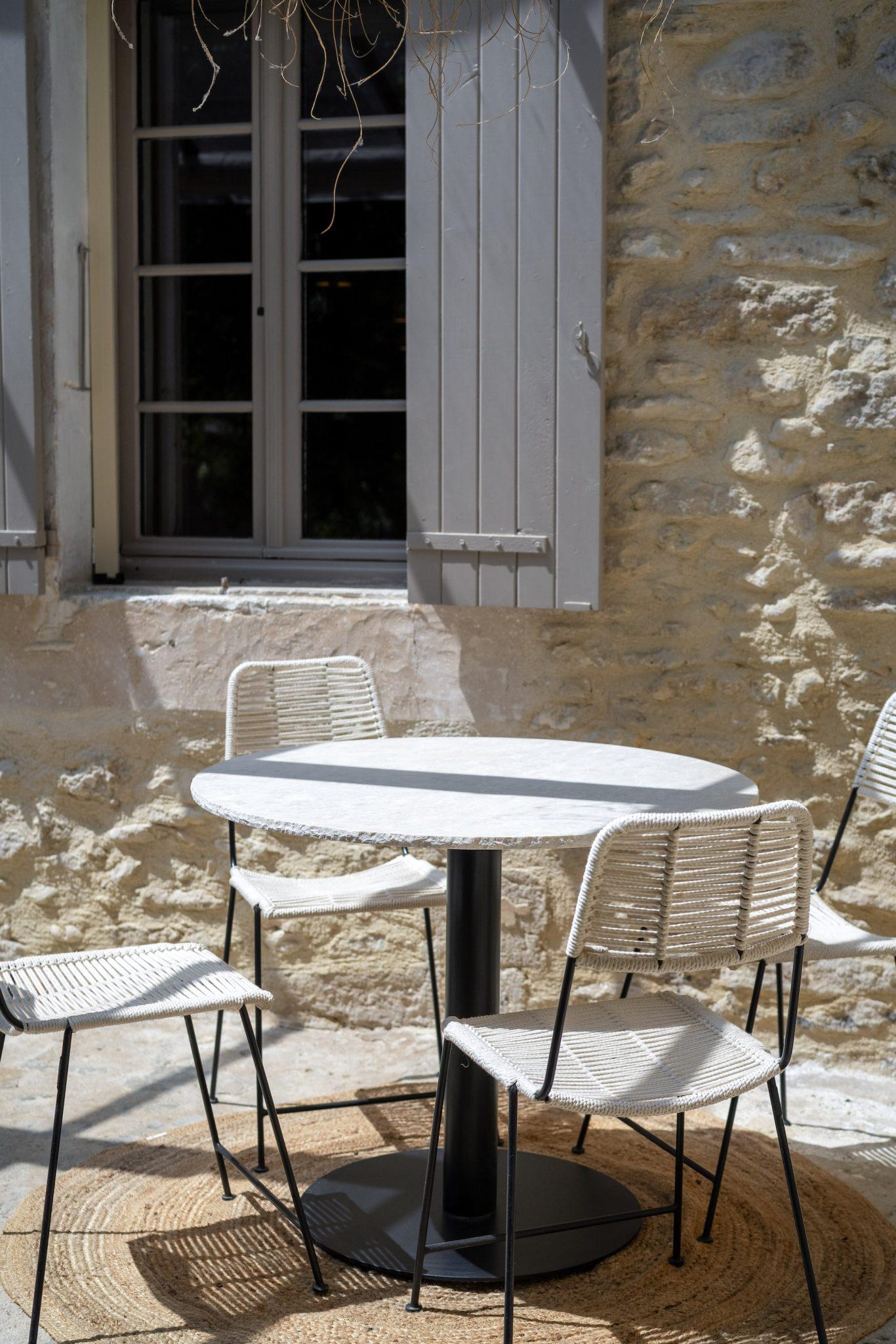 A round white table with a black pedestal is surrounded by four white woven chairs with black legs. The setting is an outdoor patio with a stone wall and a window with gray shutters in the background. Sunlight casts shadows on the floor.