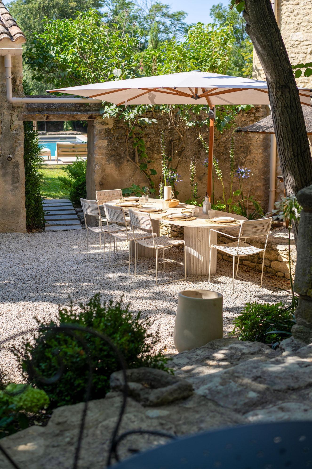 An outdoor dining area with a long wooden table and white chairs shaded by two large umbrellas. The setting is surrounded by lush vegetation and stone walls. A gravel path leads to a garden with more greenery and a glimpse of a pool in the distance.