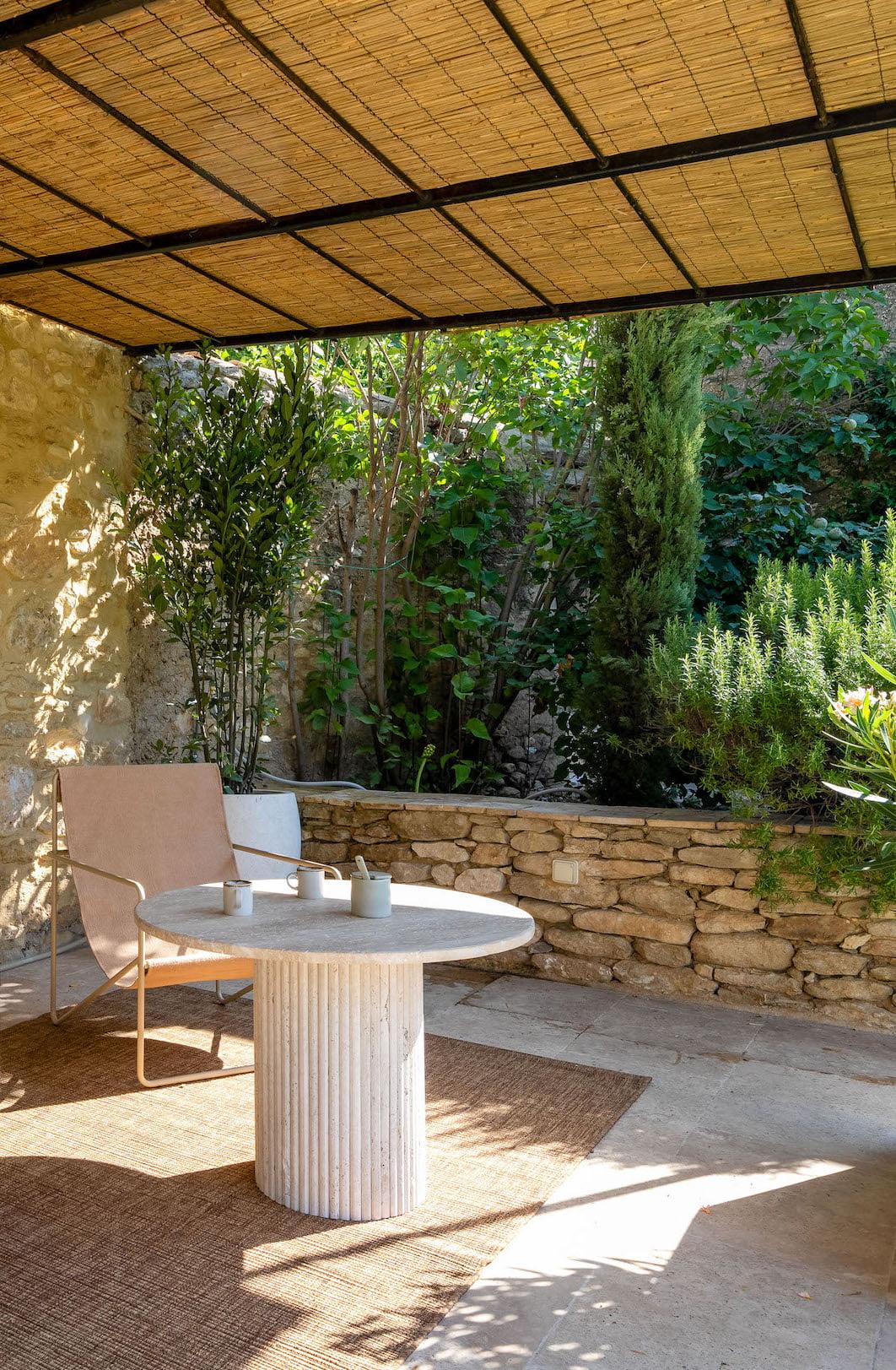 A tranquil patio features a round stone table and a minimalist chair. The space is shaded by a bamboo roof and surrounded by lush greenery and stone walls. Sunlight filters through the foliage, casting dappled light and creating a serene and welcoming atmosphere.