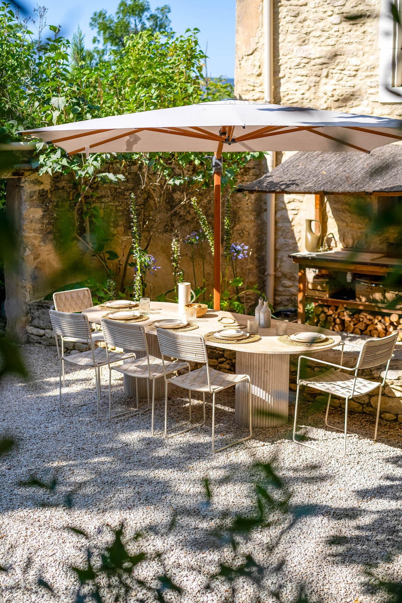 A charming outdoor dining area features a wooden table surrounded by white chairs on a gravel floor. A large umbrella provides shade. The space is adorned with greenery and backed by a rustic stone wall with stacks of firewood and garden décor.