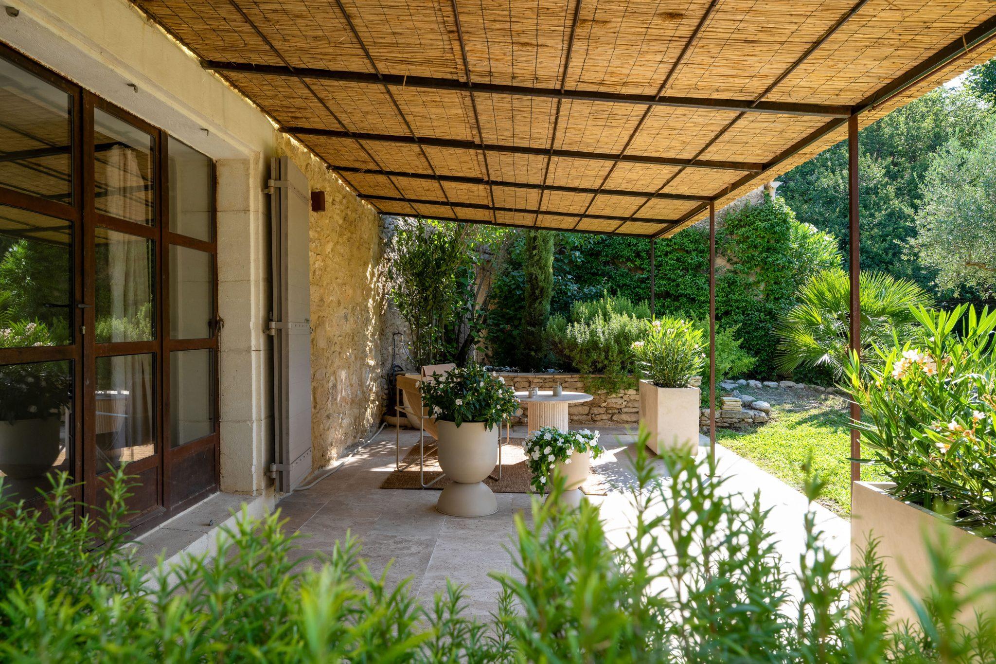 A cozy covered outdoor terrace features a dining table with chairs, surrounded by potted plants and greenery. The stone wall adds rustic charm, and the wooden roof provides shade. Sunlight filters through, creating a serene and welcoming atmosphere.